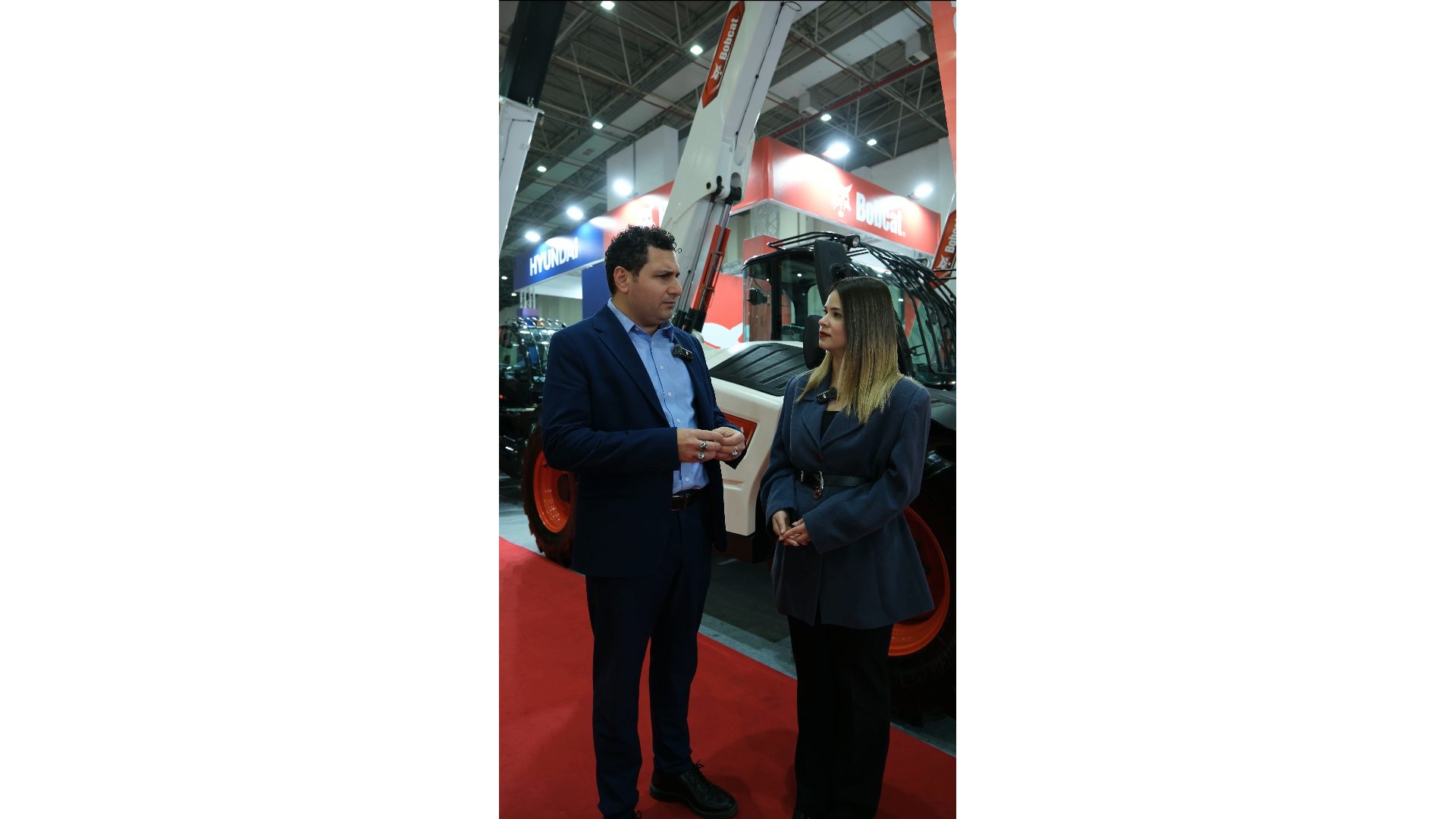 Man and woman converse at an industrial exhibition with heavy machinery in the background.