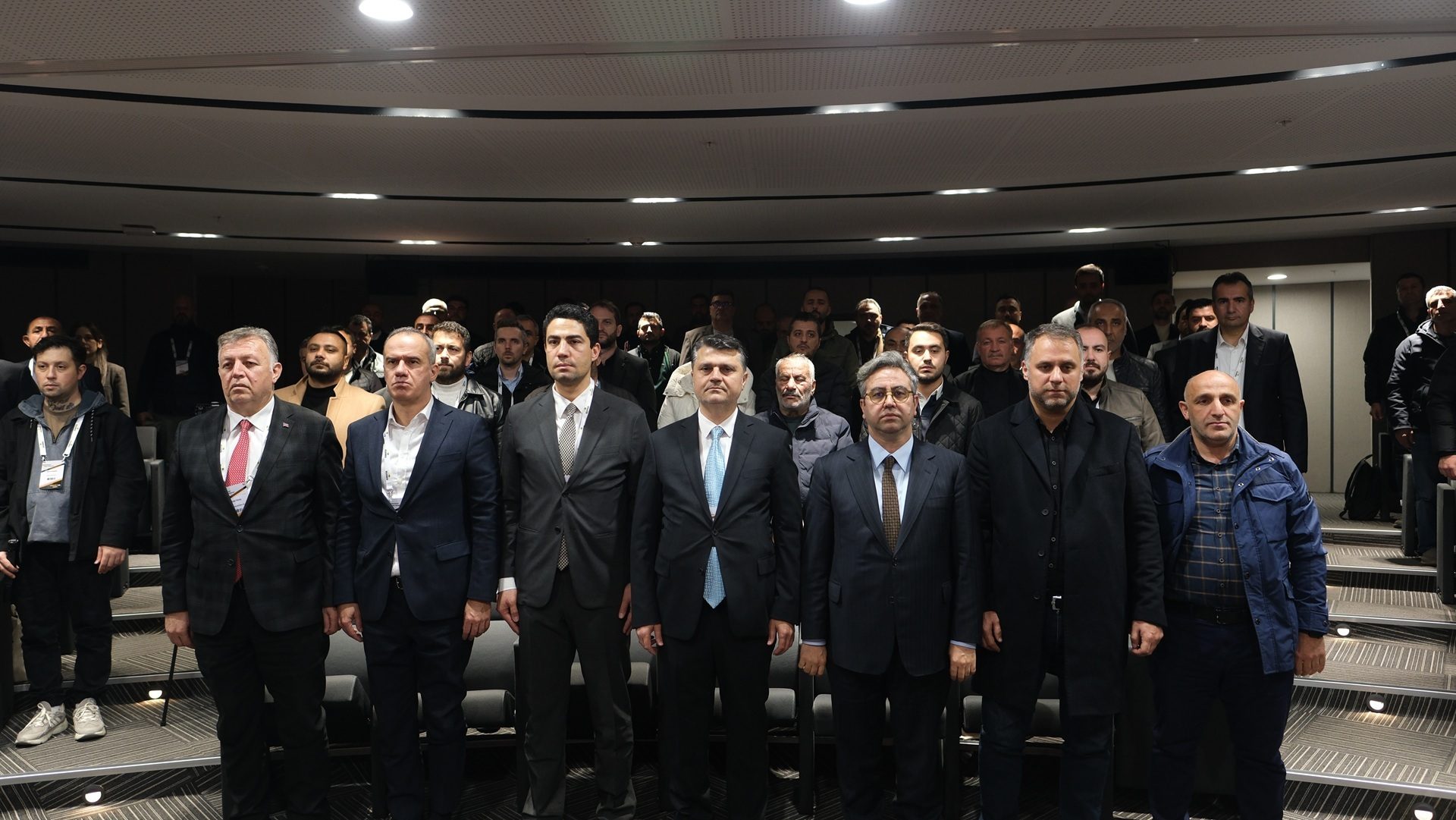 Many men, some in suits, stand in rows in a room, appearing attentive.