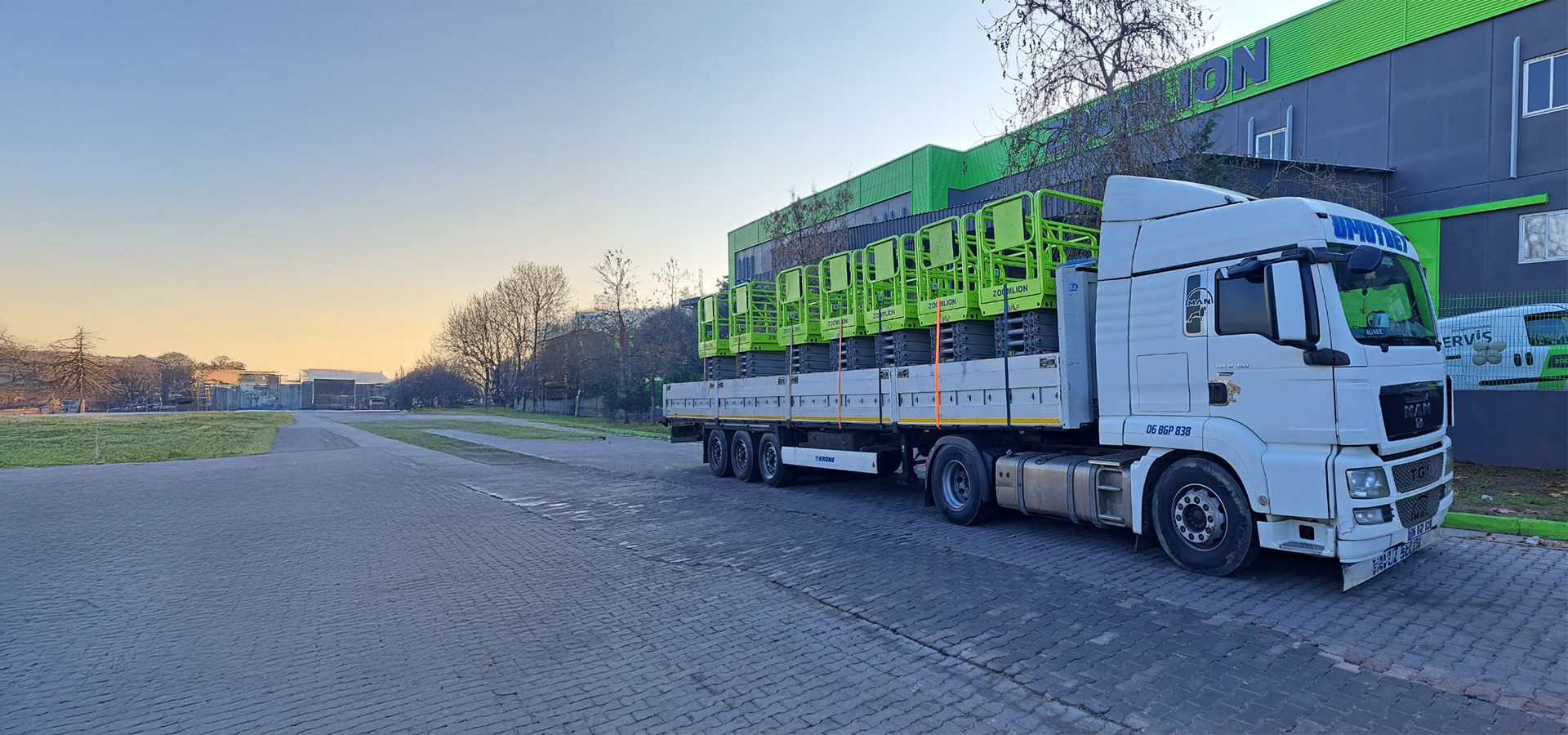 White truck carrying multiple green scissor lifts on a flatbed trailer near a building.