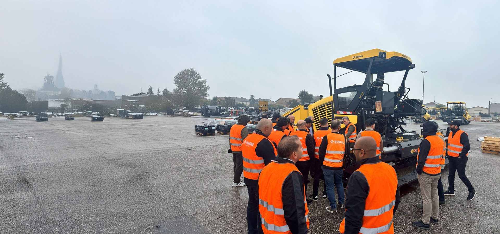 Men in orange safety vests gathered around a yellow construction machine outdoors on a hazy, cold day.