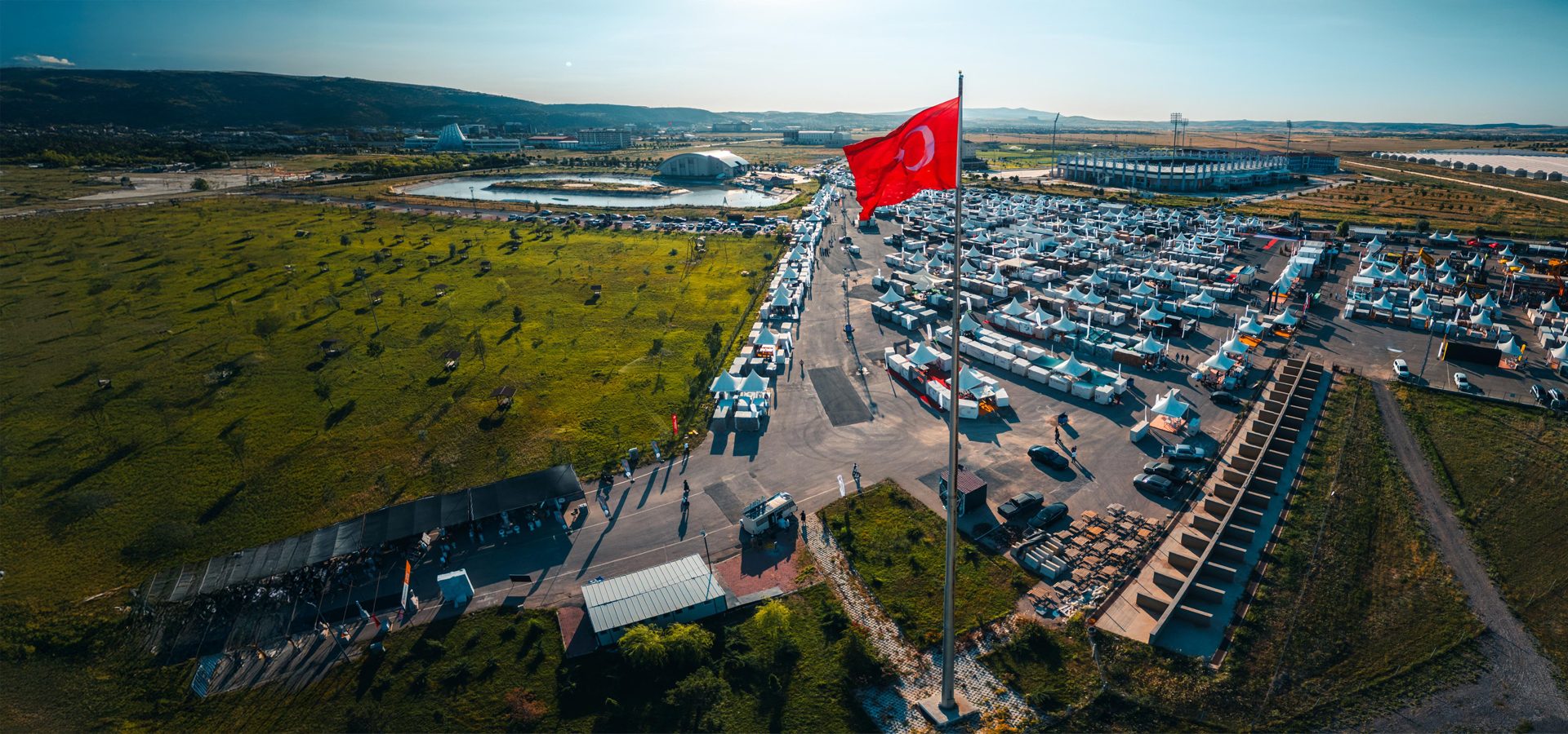 Aerial: Huge Turkish flag dominates an outdoor fair with rows of white tents & stadium.