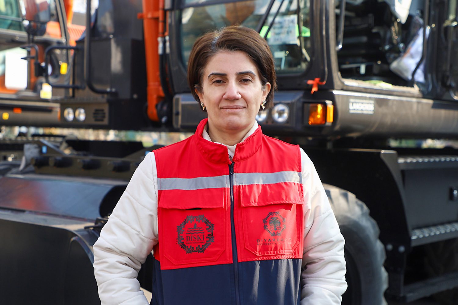 Woman in a work vest standing before heavy machinery.