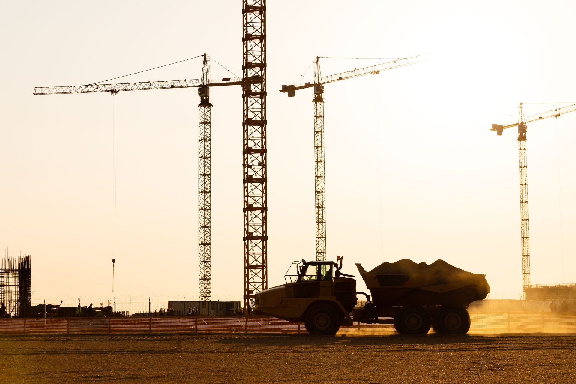 Silhouetted construction site with cranes and a dump truck against a bright, hazy sky.