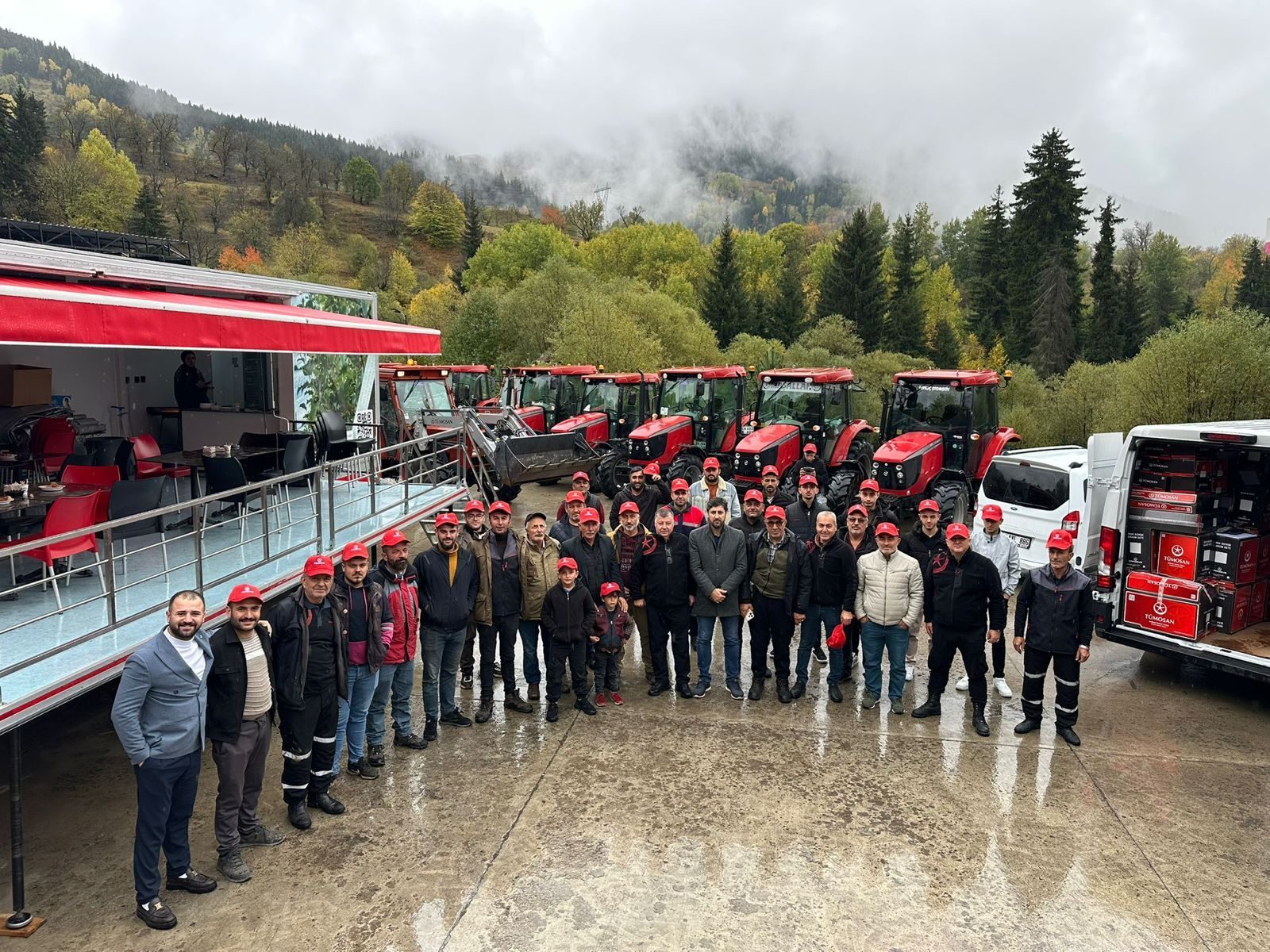 Group of men in red caps pose with red tractors, a loaded van, and a misty mountain forest background.