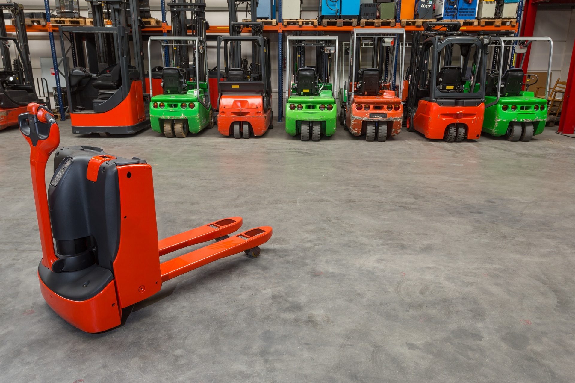 Orange electric pallet jack in a warehouse with a line of red and green forklifts behind it.