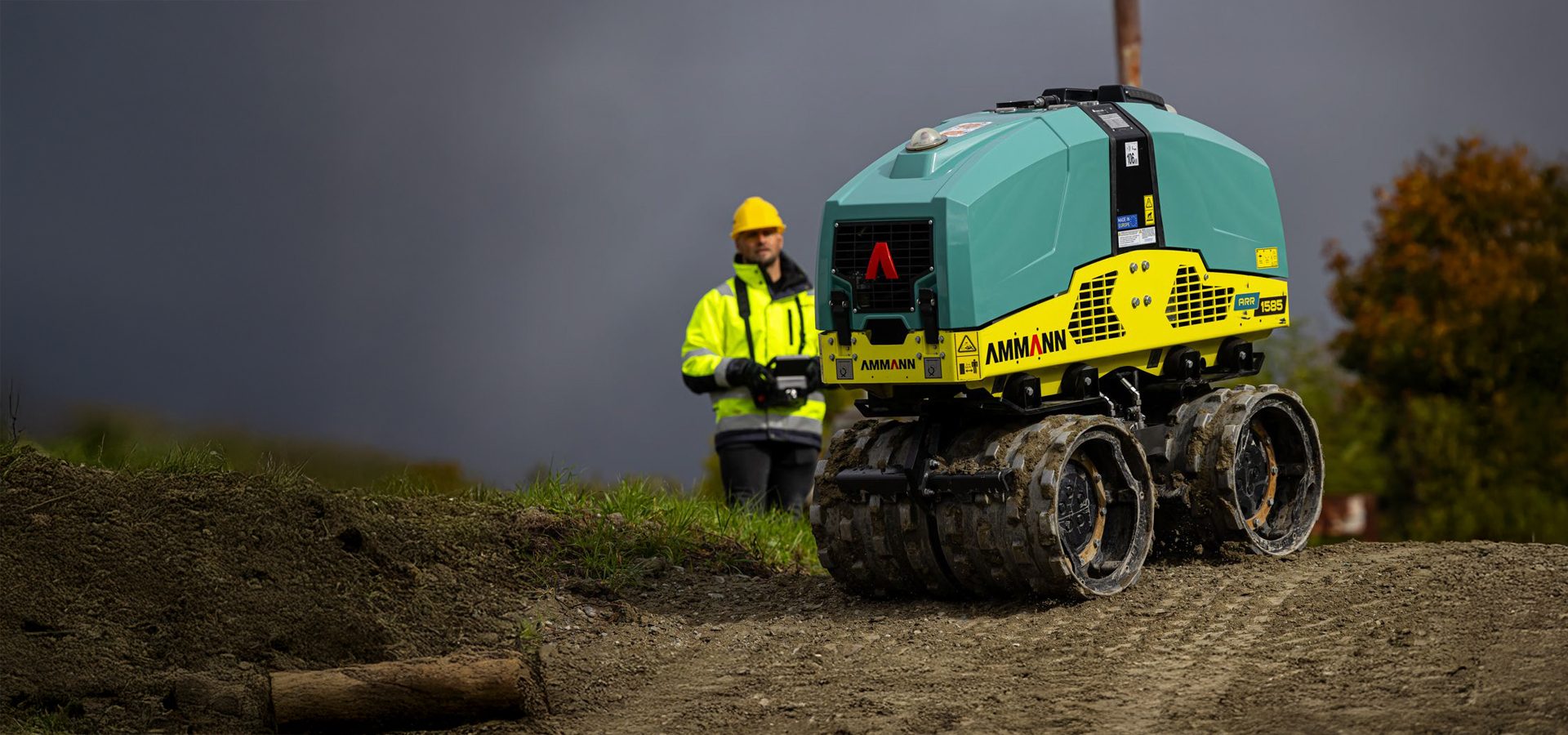 Construction worker remotely operates an Ammann soil compactor on a dirt path.