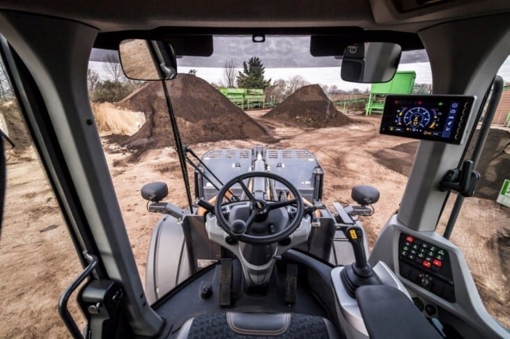 Inside a wheel loader cab: steering wheel, digital display, and a view of dirt piles.