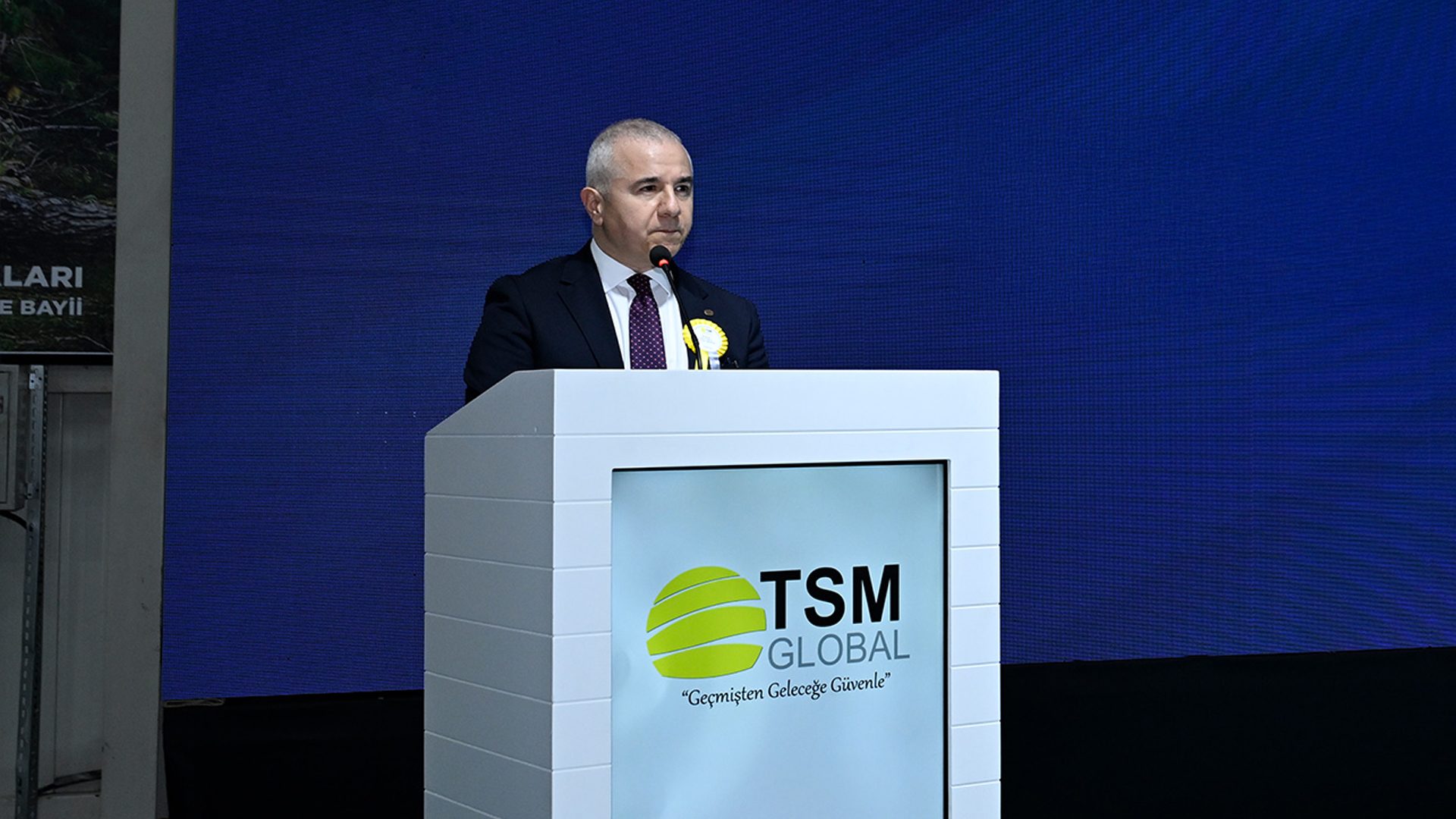 A man in a suit with a yellow rosette speaks at a white podium displaying the "TSM GLOBAL" logo against a blue background.