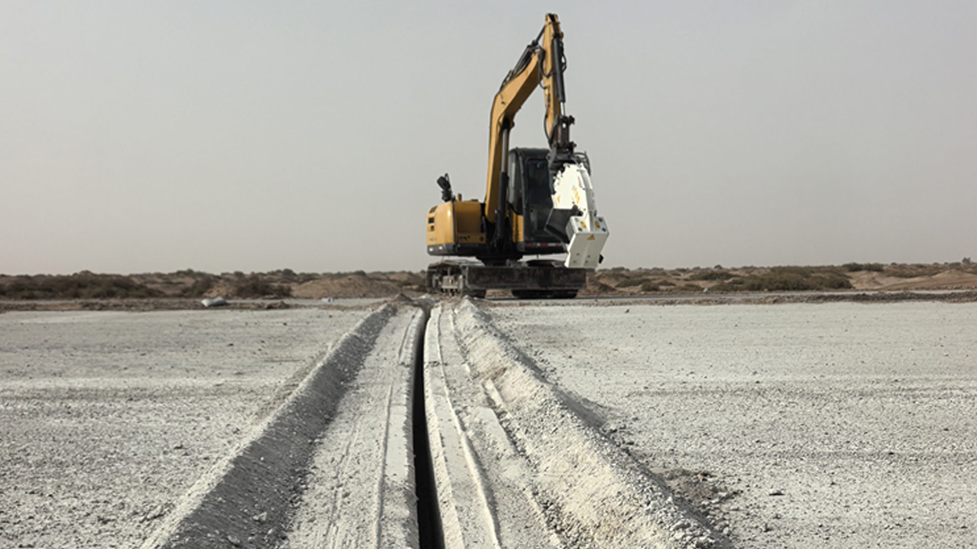 Yellow excavator with trenching attachment digging a long, straight trench in arid ground.