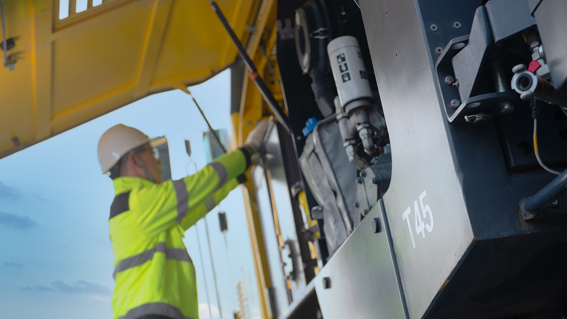 Worker in hard hat inspecting heavy machinery's open engine compartment, marked 'T45'.