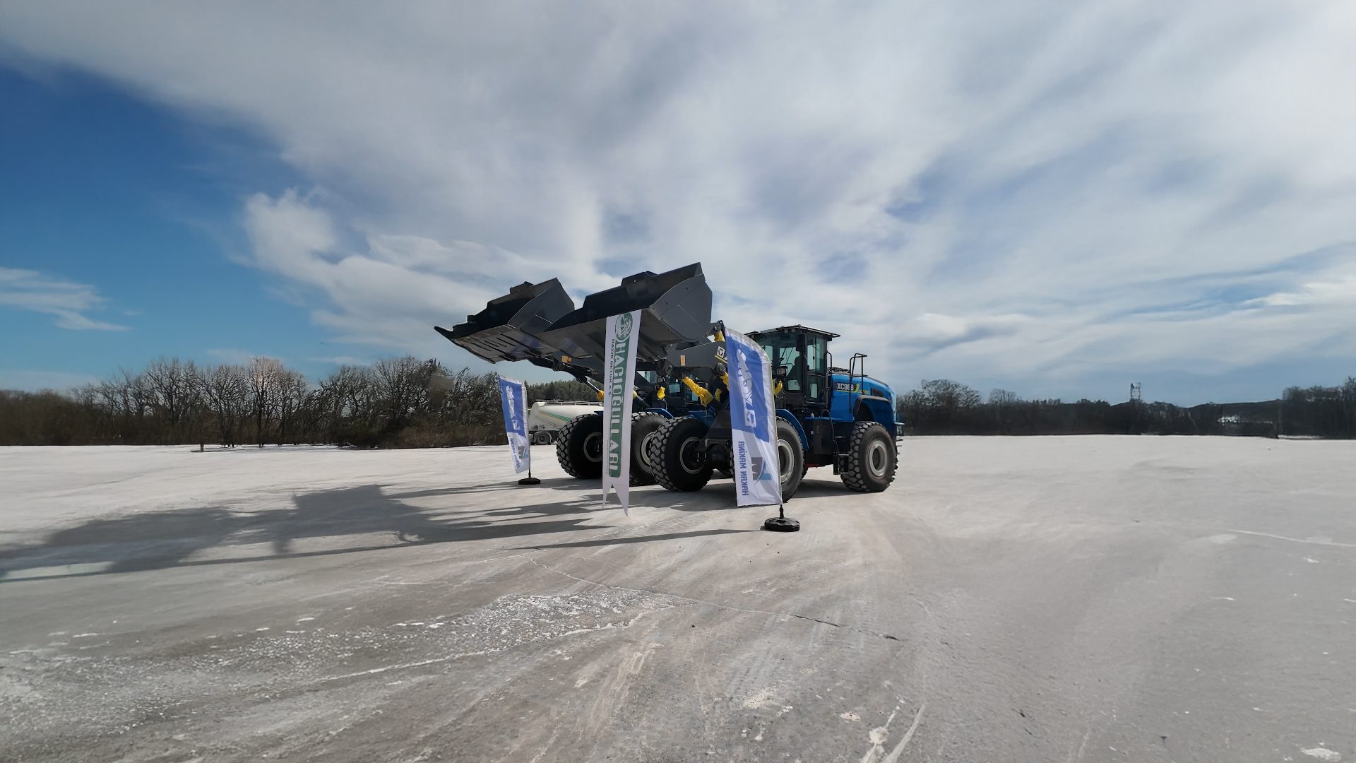 Two blue wheel loaders with raised buckets on a salt flat. Flags, cloudy sky, and bare trees.