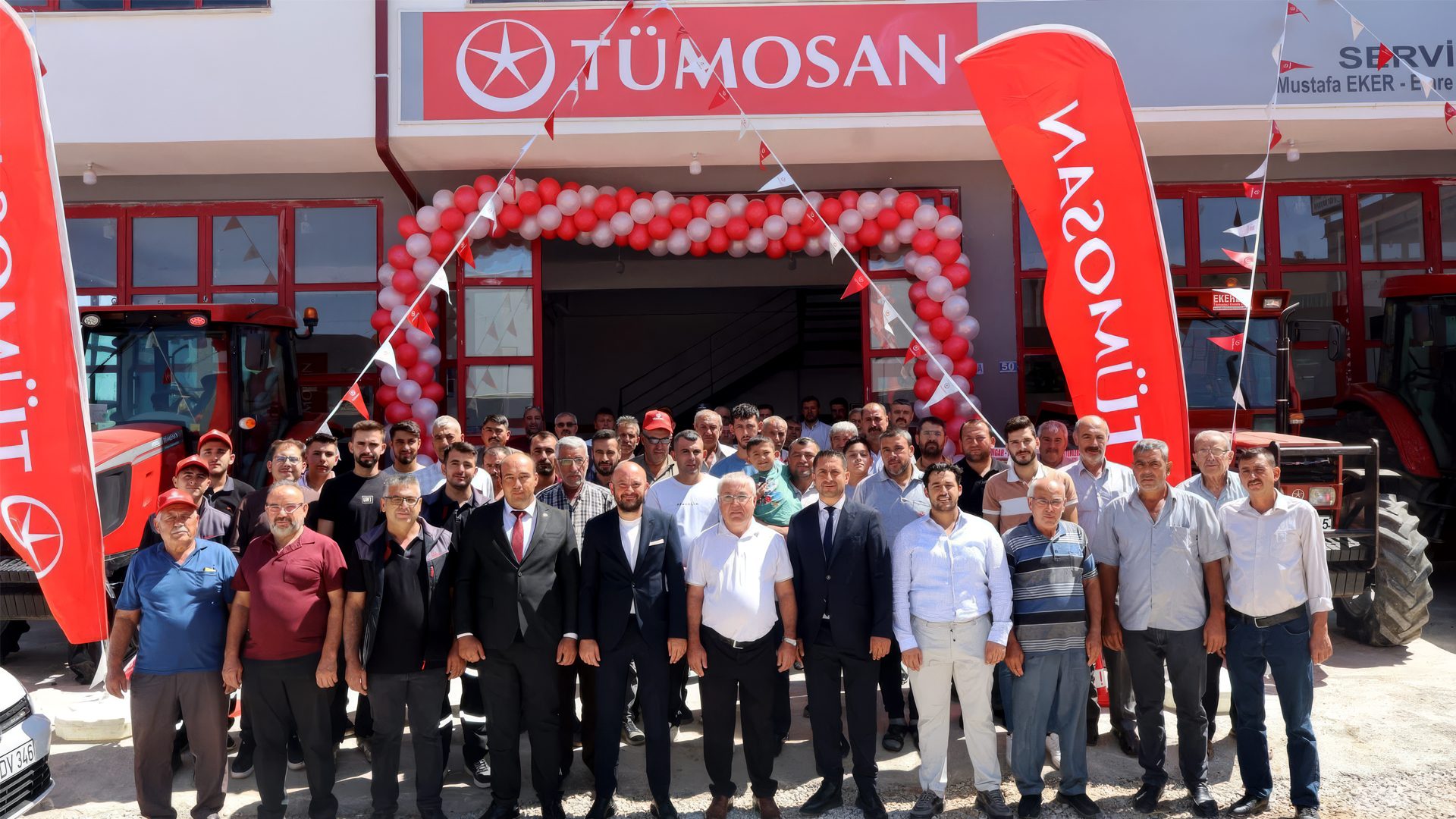 A large group of men pose at the entrance of a red TÜMOSAN building decorated with balloons and tractors.