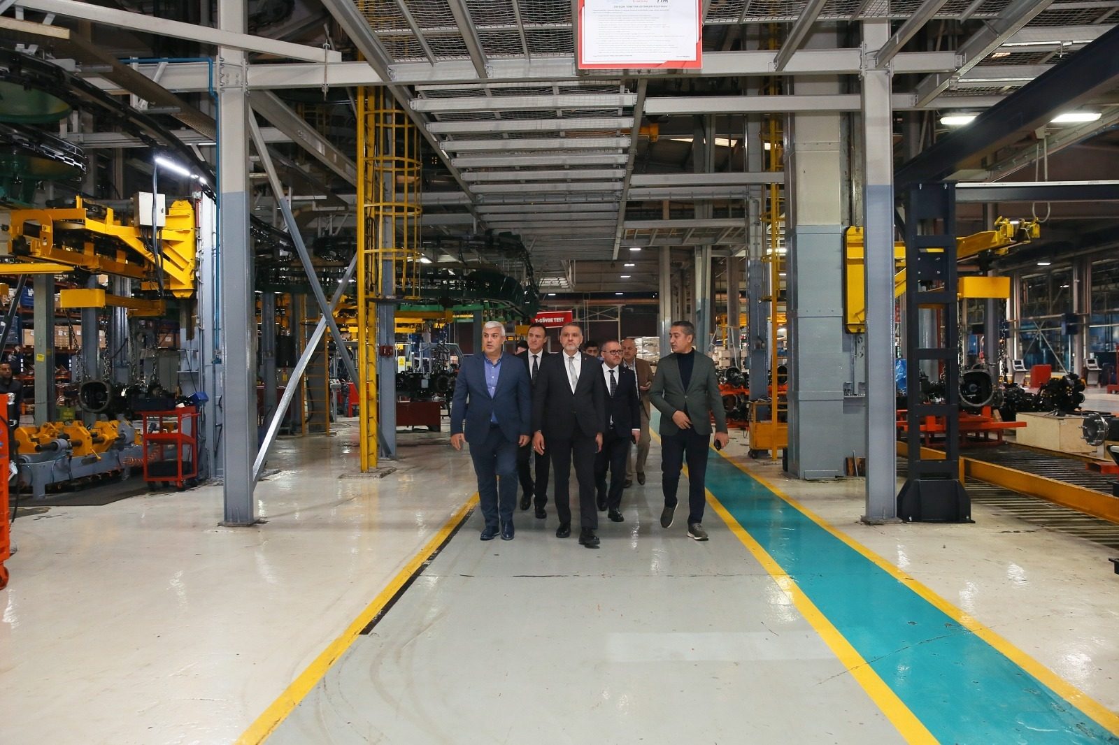 A group of men in suits walk down a designated aisle in a busy factory, surrounded by machinery.