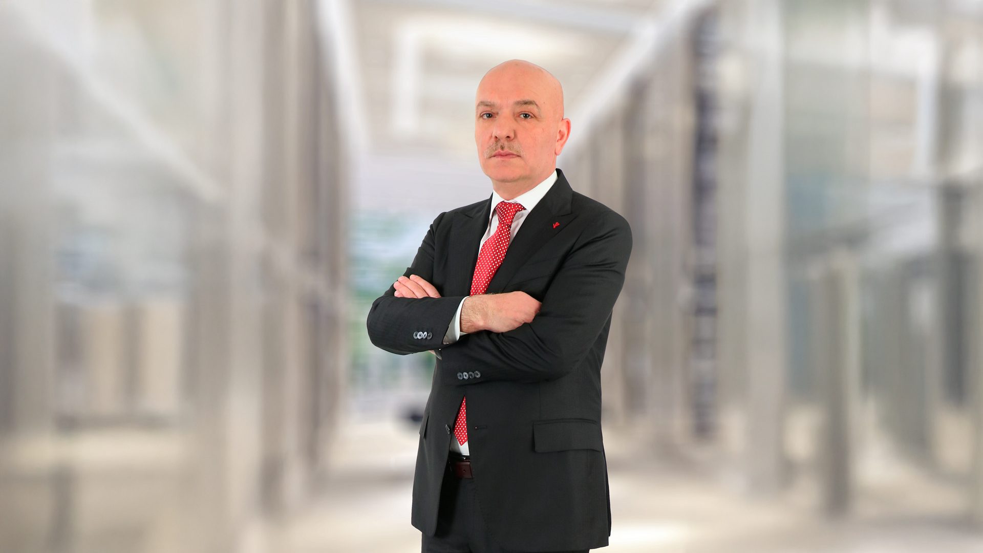 Bald man with a mustache, in a suit and red tie, arms crossed, against a blurred office background.