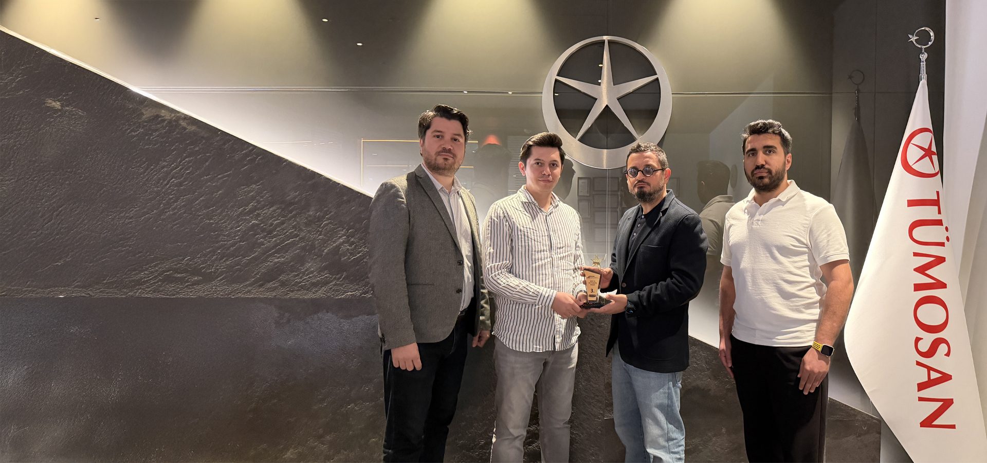 Four men, two holding a small award, stand in an office with a star logo and a TÜMOSAN flag.