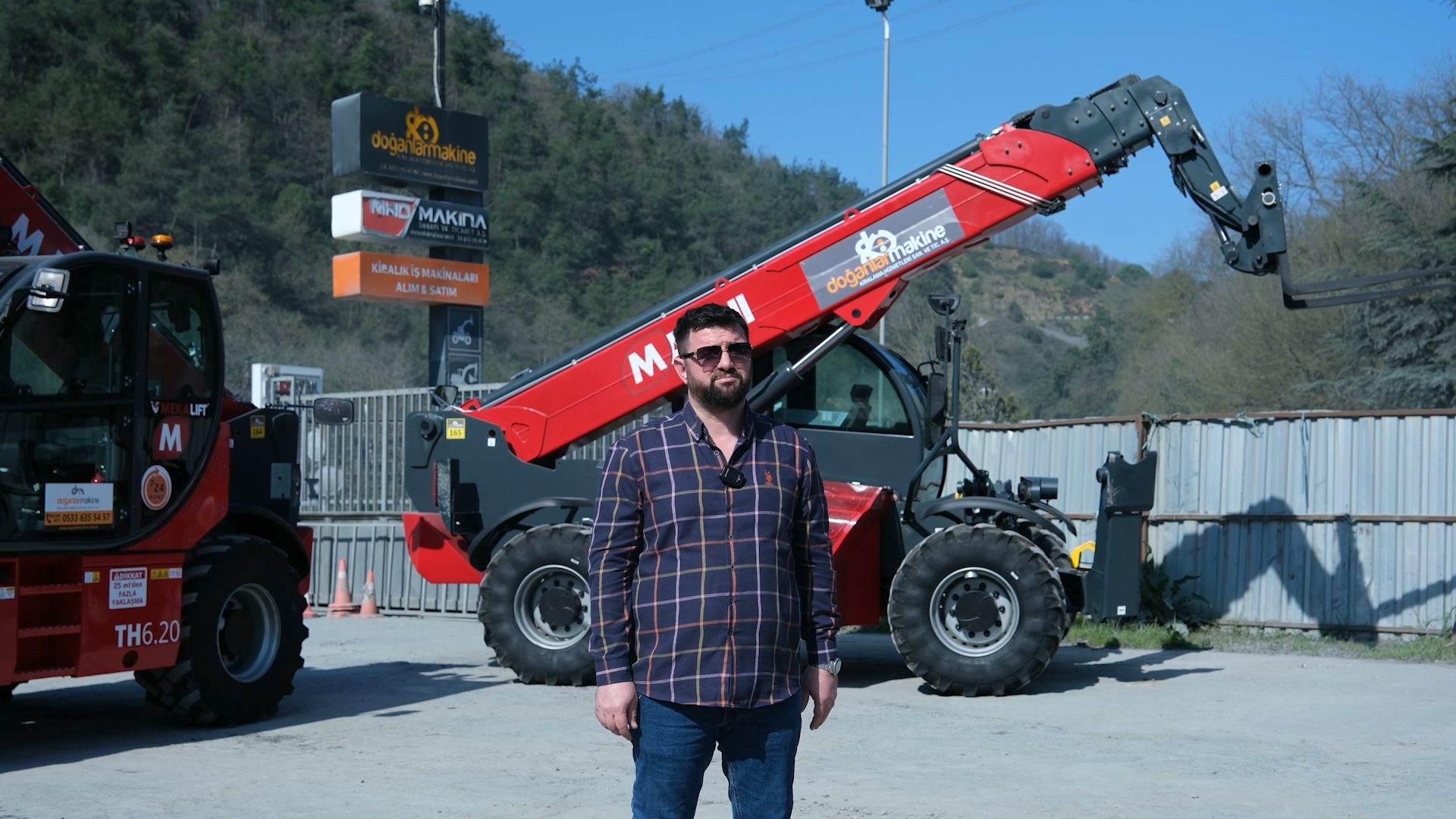 Man in sunglasses standing before two red telehandlers and a business sign under a clear sky.