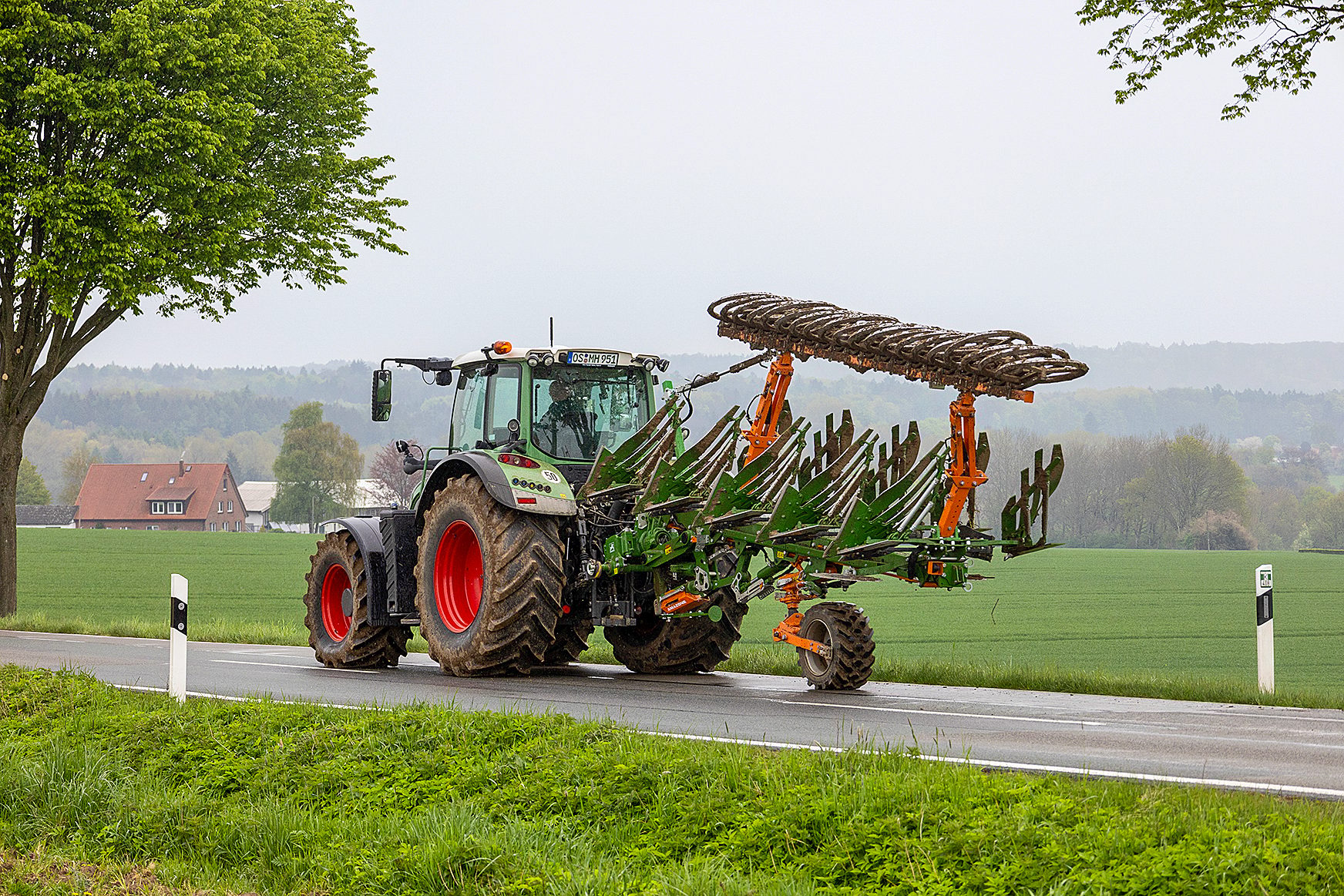 Green tractor pulling a large multi-furrow plow on a rural road with fields and trees.