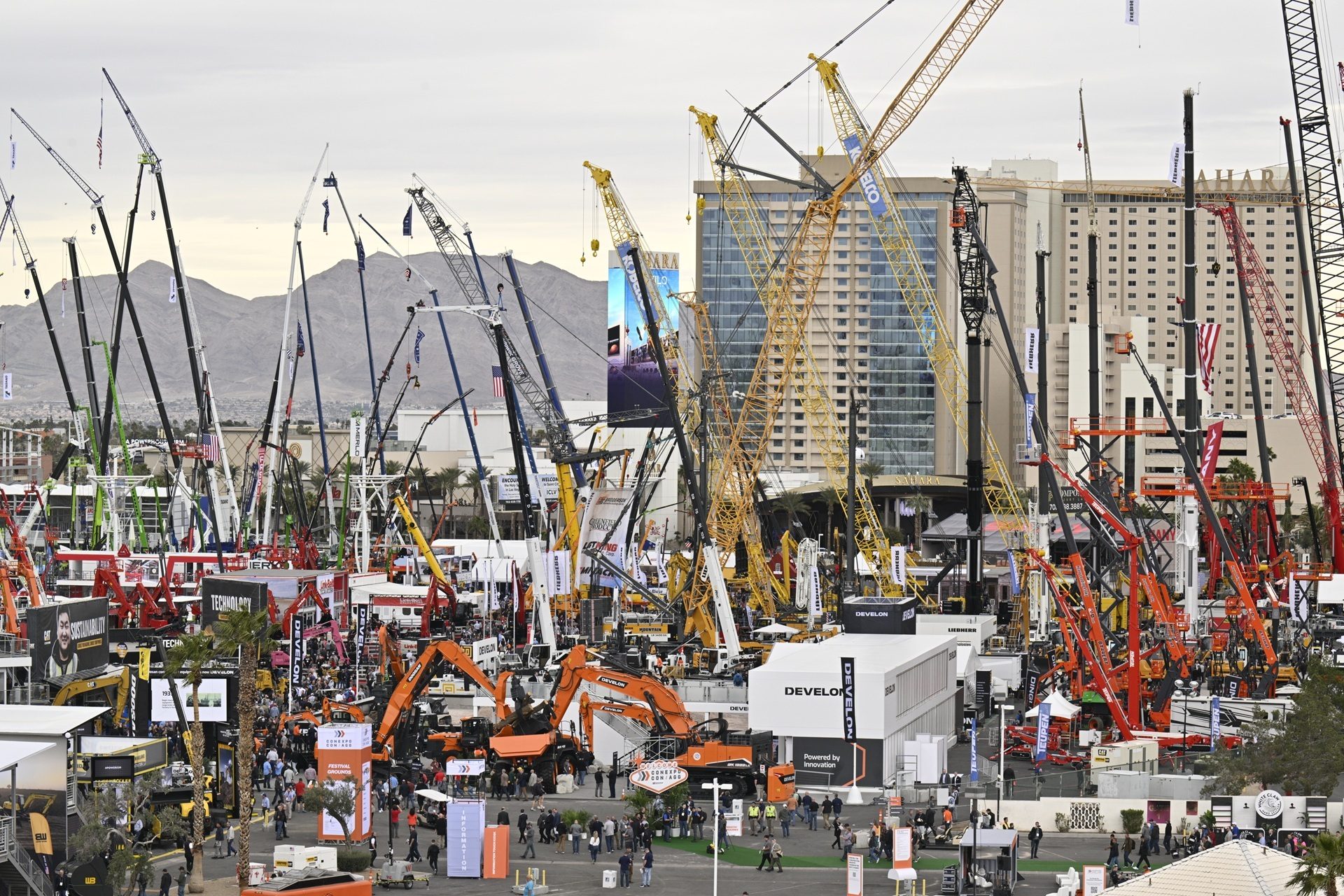 Outdoor construction trade show filled with numerous cranes, heavy machinery, people, and buildings.