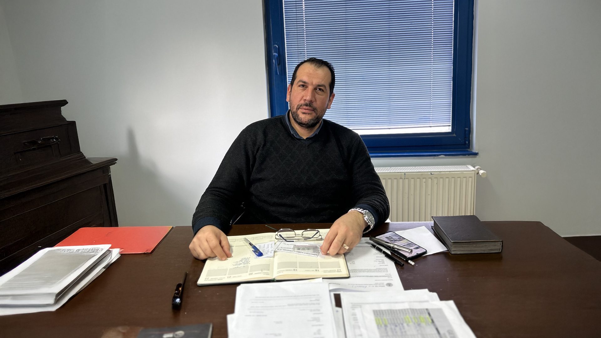 Bearded man in black sweater sits at desk with open planner, looking at camera.