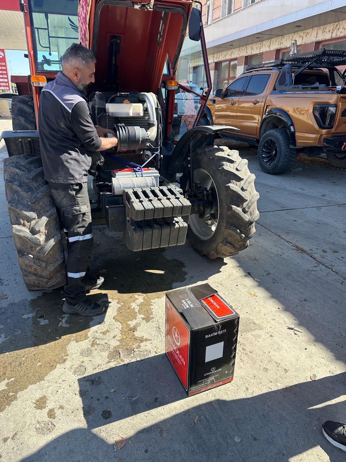 A man works on a tractor engine, with a TÜMOSAN parts box on the ground next to him.