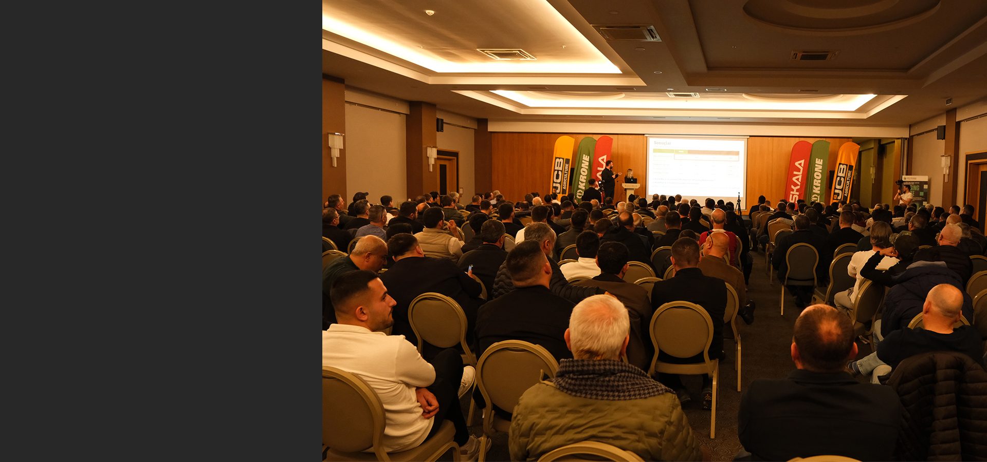 Audience of men watching a presentation with JCB, Krone, Skala banners on a stage.