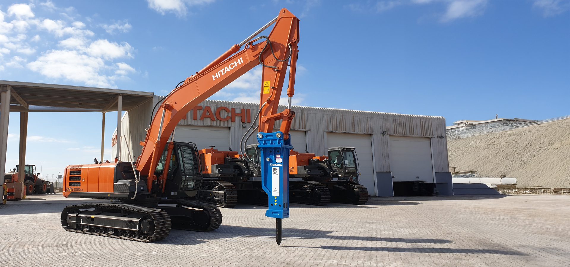 An orange Hitachi excavator with a blue hydraulic breaker attachment parked outside a building on a sunny day.
