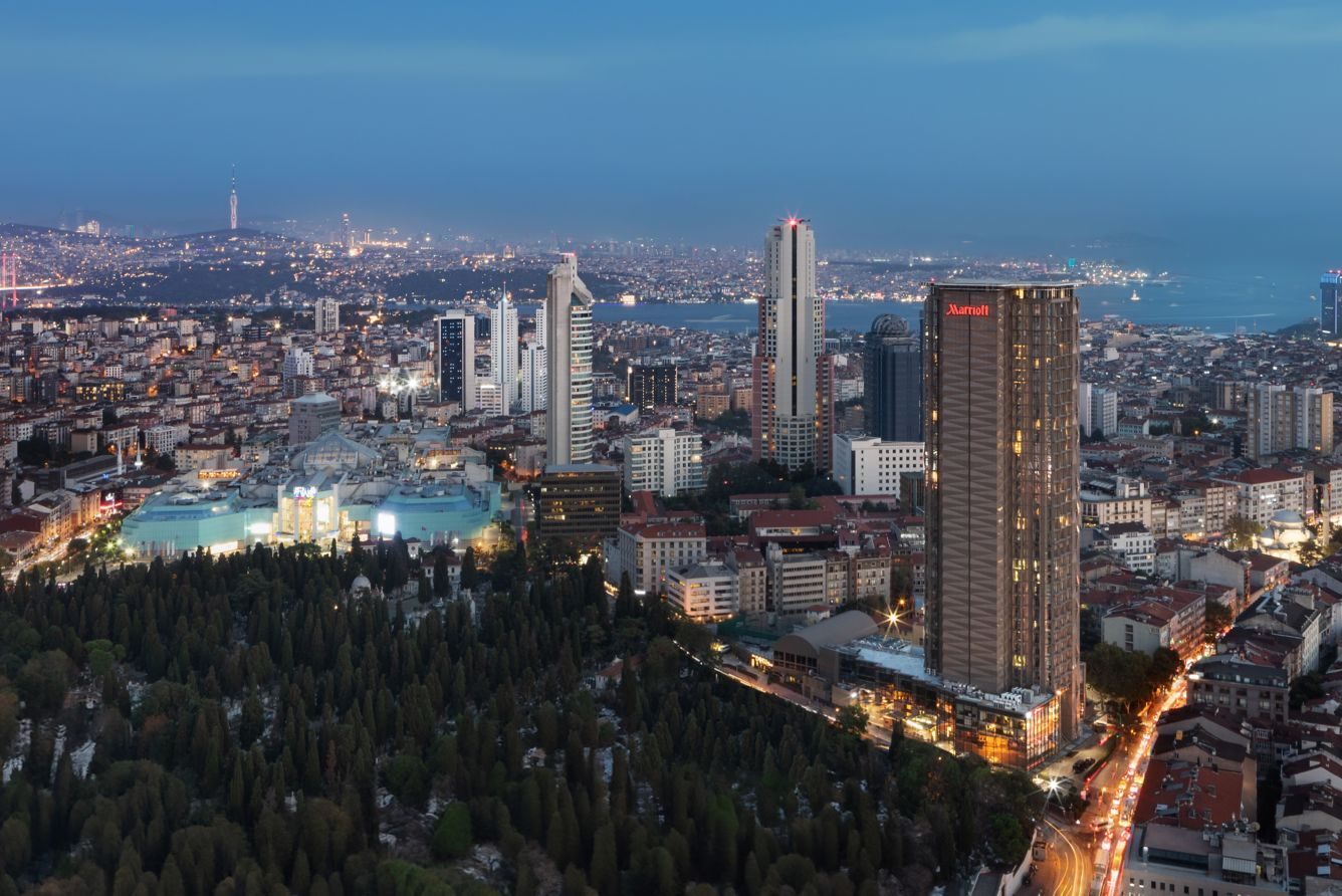 Aerial view of a city at dusk, with illuminated buildings, a dense forest, and a Marriott tower.