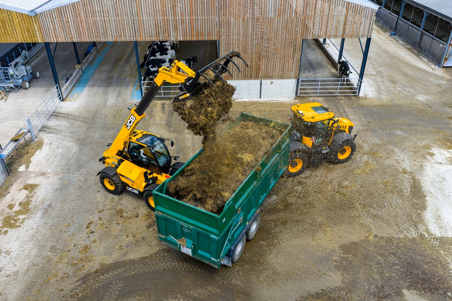 Yellow JCB telehandler loading farm material into a green trailer pulled by a tractor on a farm.