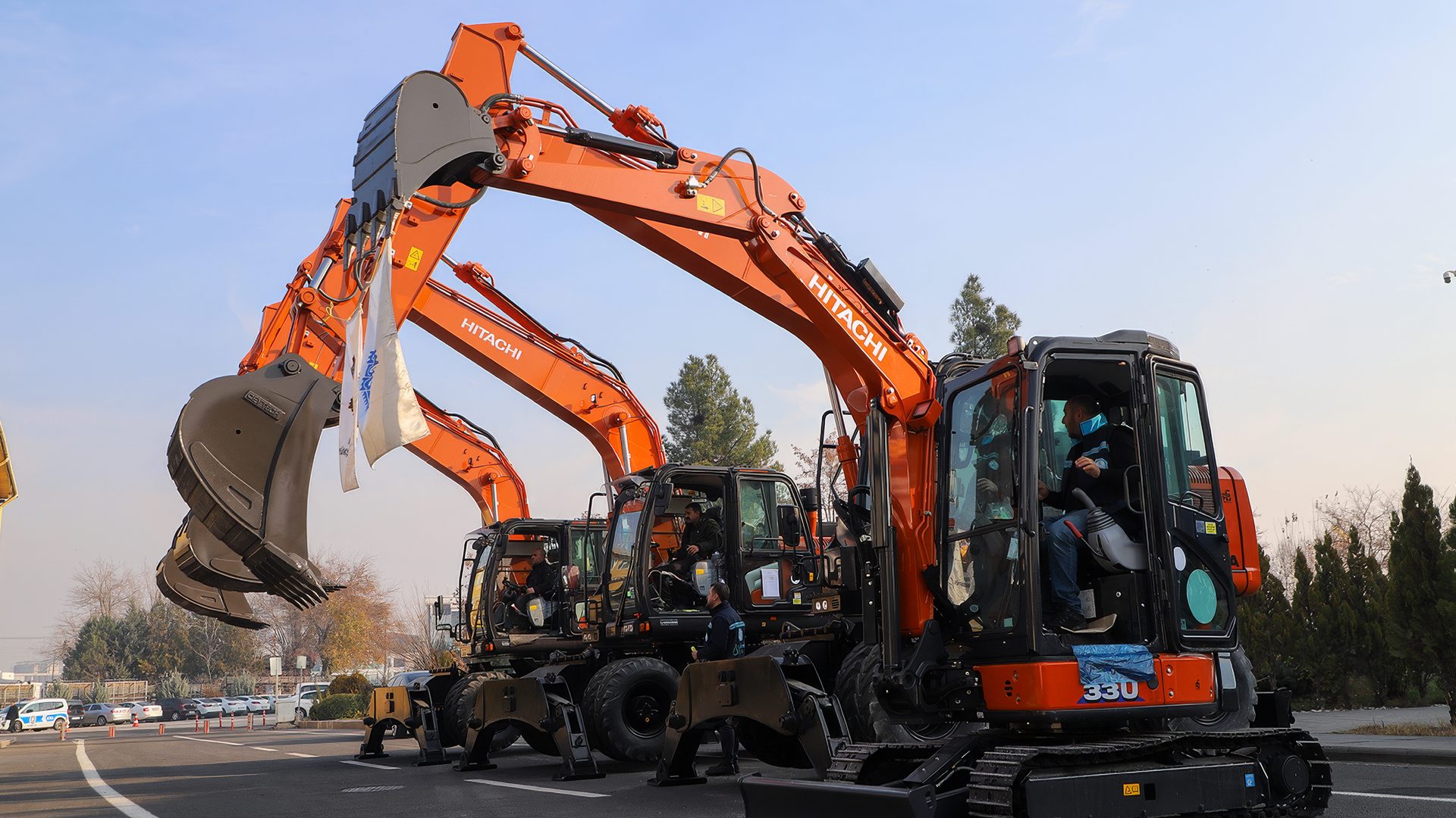 Multiple orange Hitachi excavators lined up with people in their cabs.