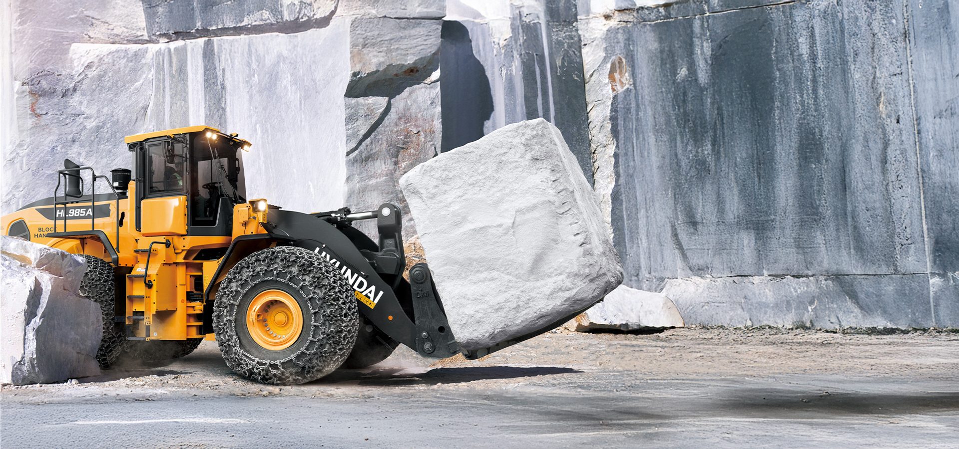 A yellow Hyundai wheel loader with chained tires lifts a huge white stone block in a quarry.