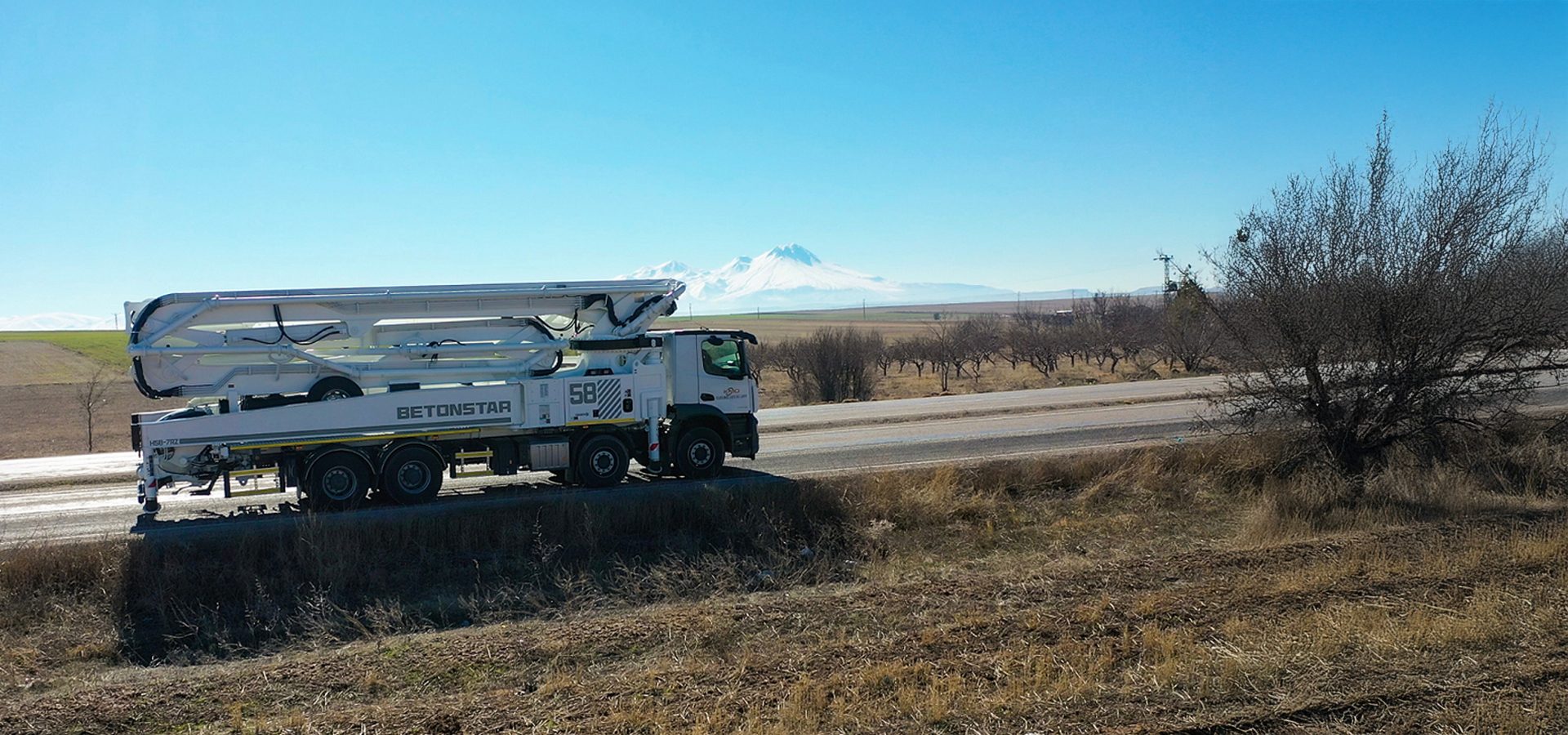 A white Betonstar concrete pump truck travels on a road with snow-capped mountains and fields under a clear sky.