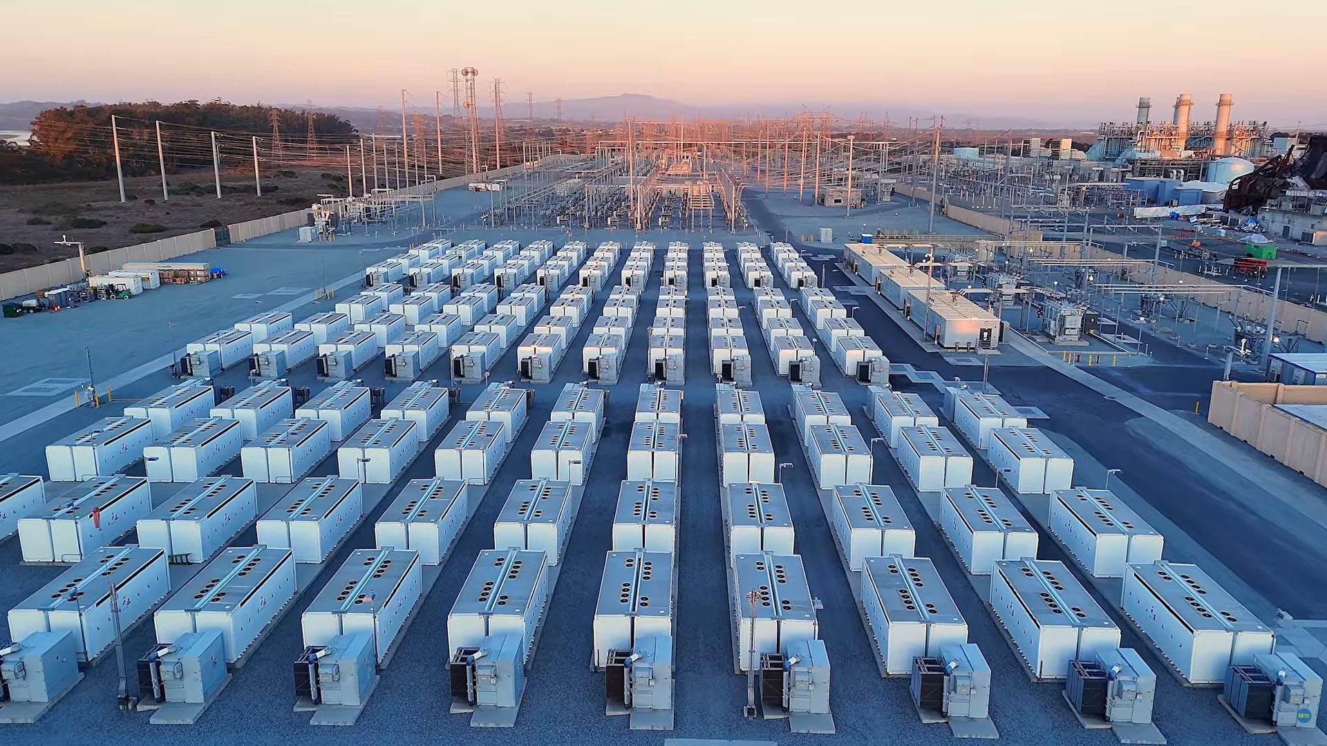 Aerial view of a large battery energy storage facility with rows of white containers, a substation, and power plant.