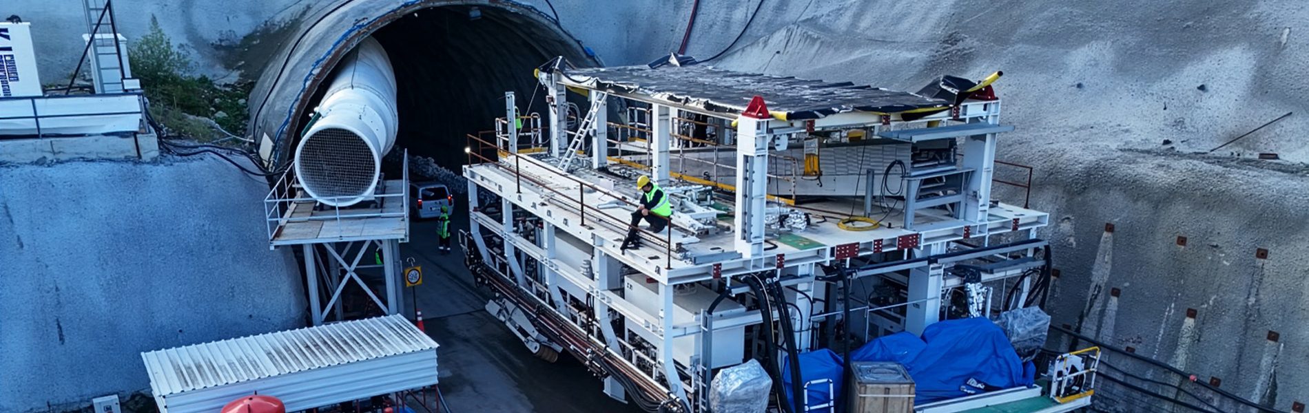 Tunnel construction site at an entrance with large machinery, a ventilation pipe, and workers.