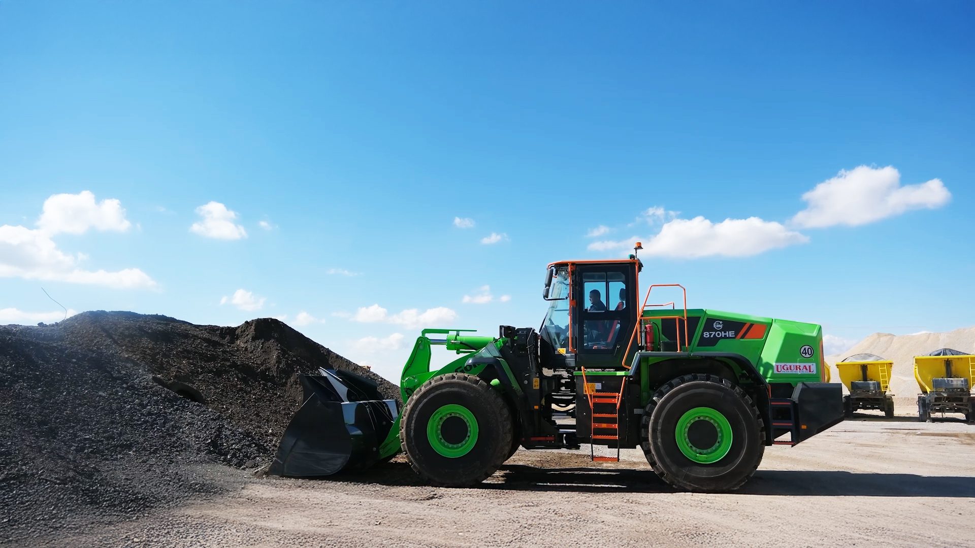 Green construction loader with dark material pile, blue sky.