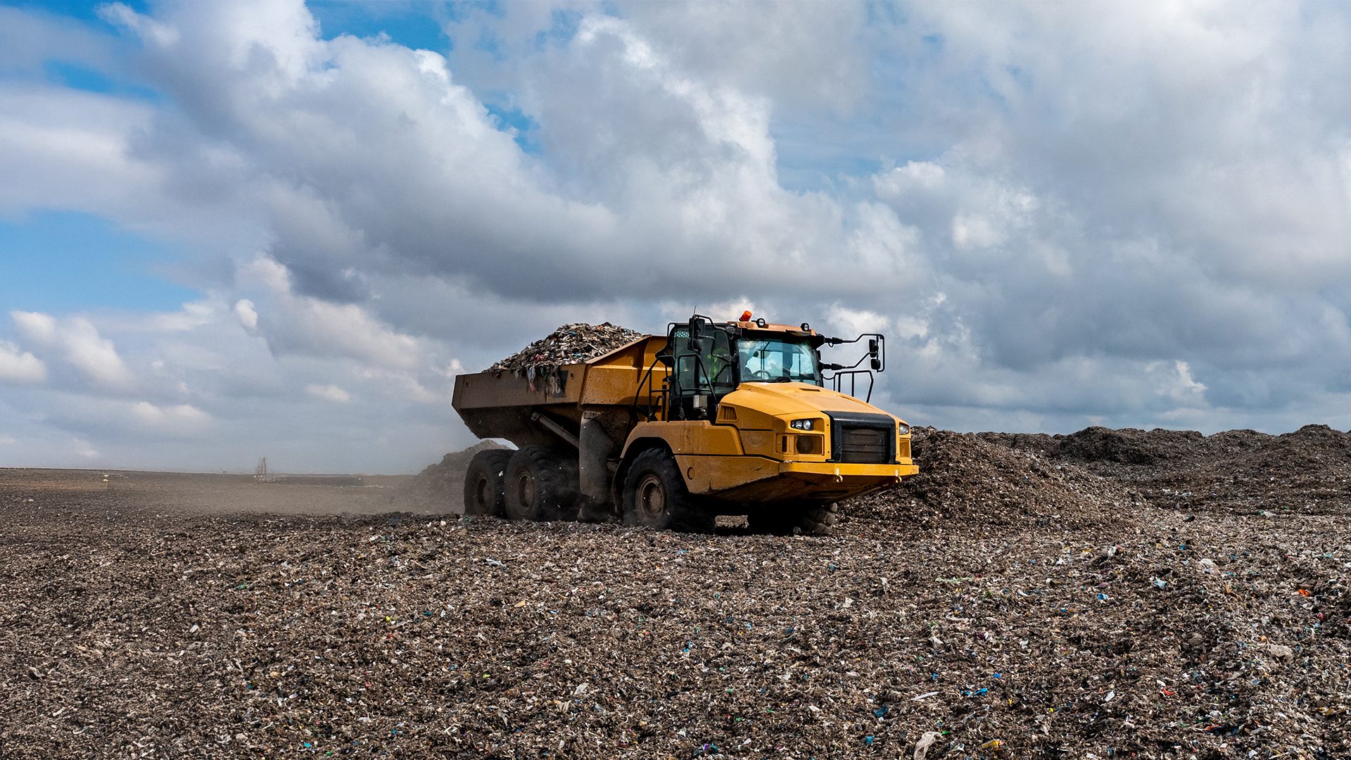 Large yellow dump truck filled with waste at a landfill under a cloudy sky.