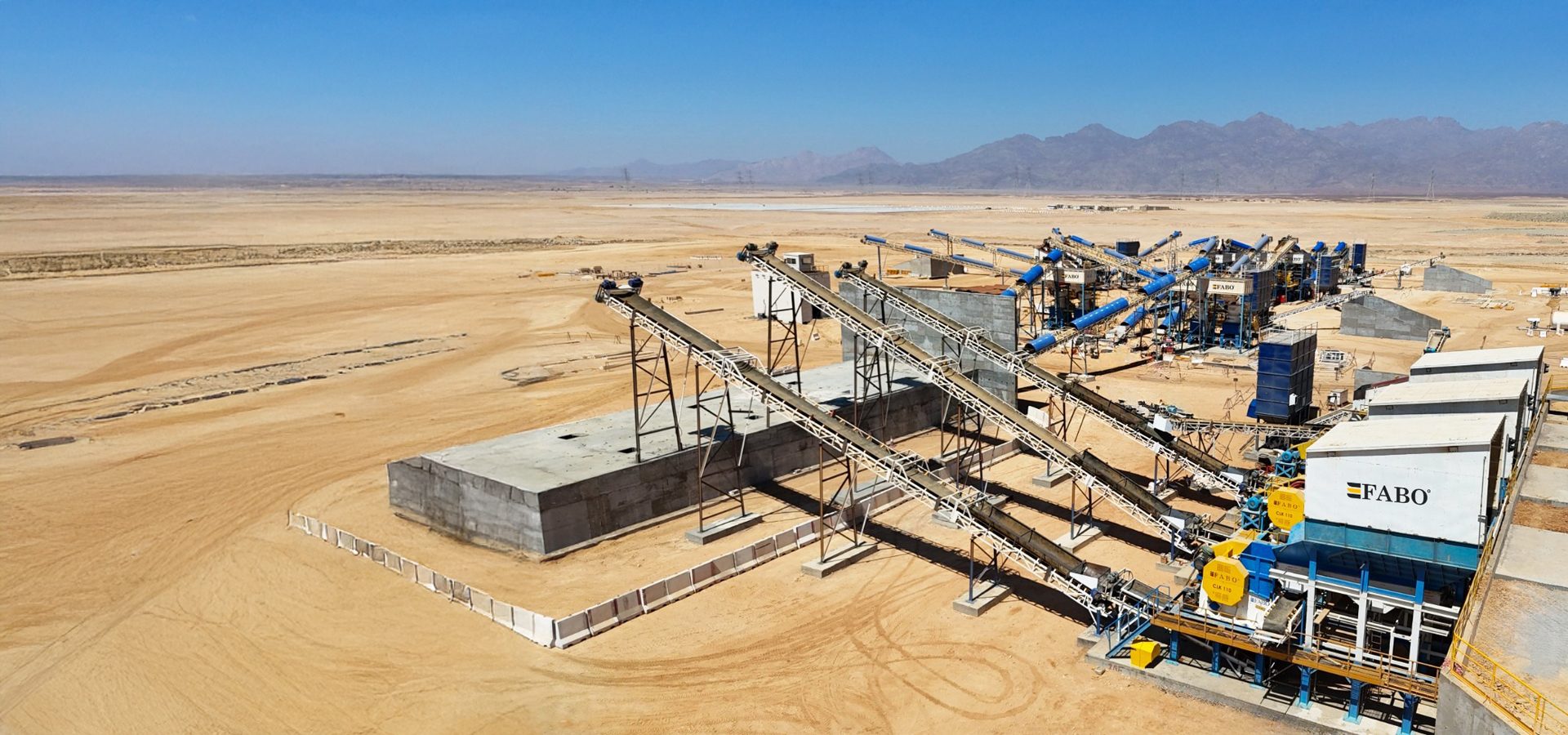 FABO rock crushing plant with conveyor belts in a desert, under a clear sky.