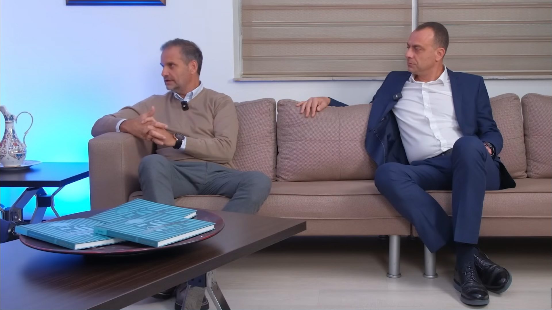 Two men sit on a couch in a casual interview or conversation setting, with books on a table.
