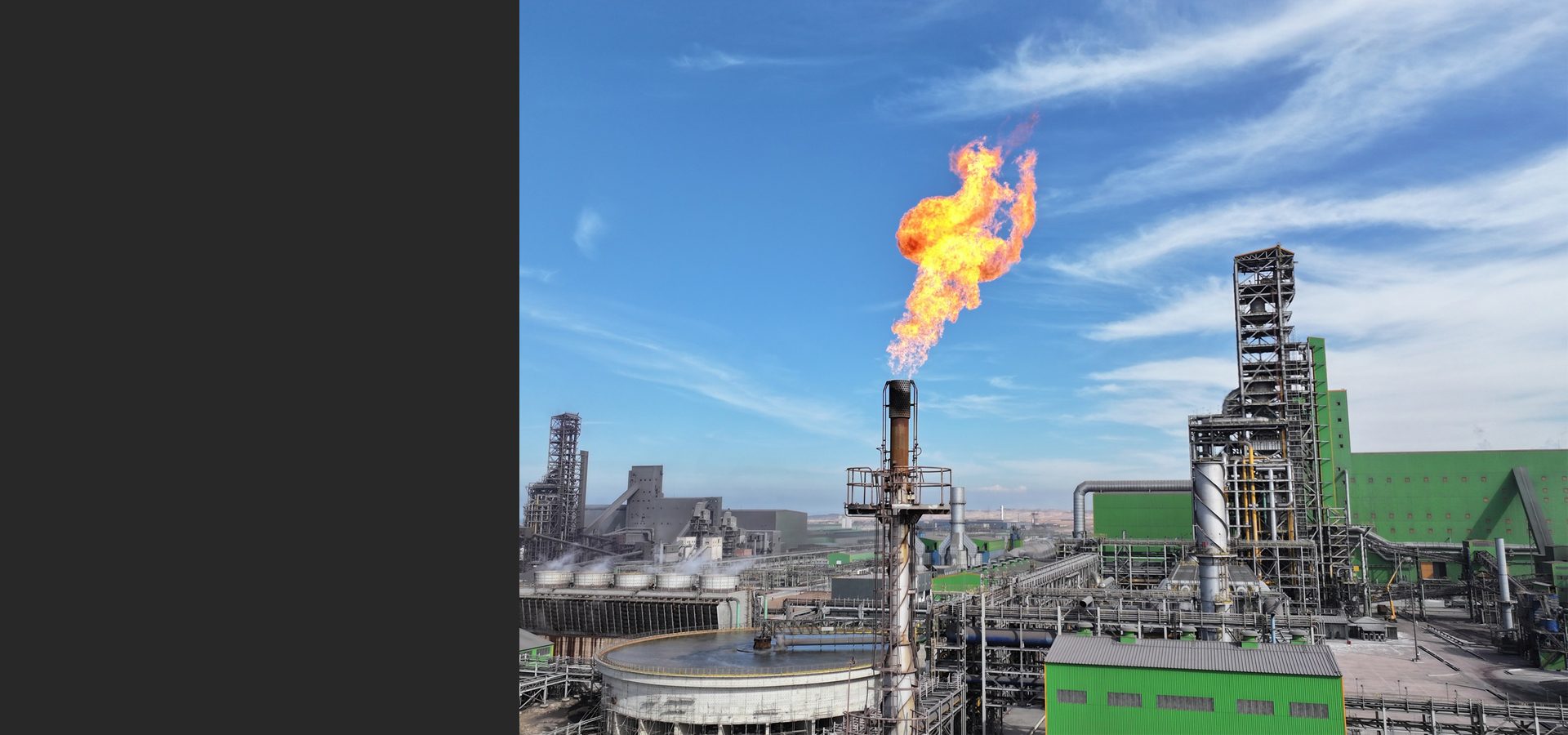 An industrial flare burns brightly against a blue sky, towering over a vast complex of industrial structures.