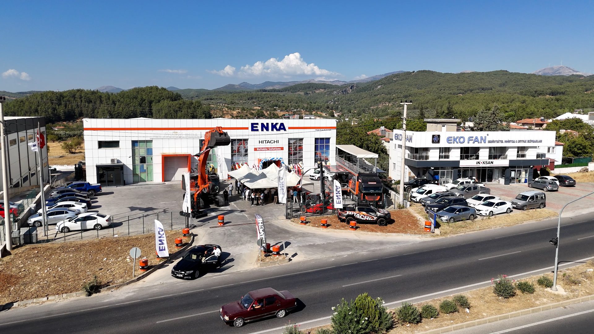 ENKA HITACHI and EKO FAN buildings, with machinery, cars, and a road. Sunny day, forested hills.