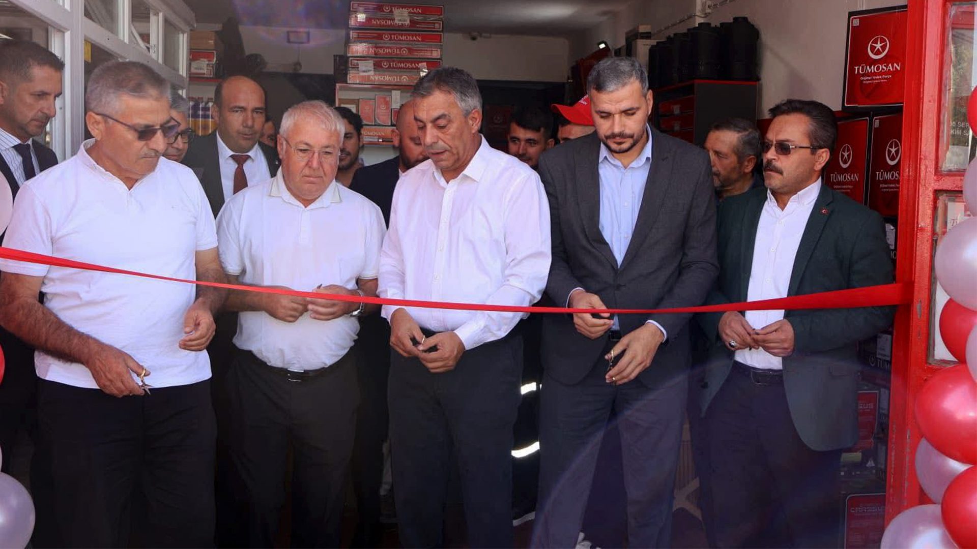 Several men in formal and semi-formal attire cutting a red ribbon at a TÜMOSAN store opening.