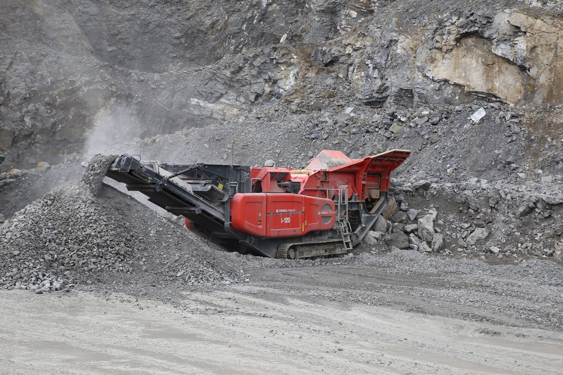 A red Terex Finlay I-120 rock crusher operates in a quarry, forming a pile of crushed gravel.