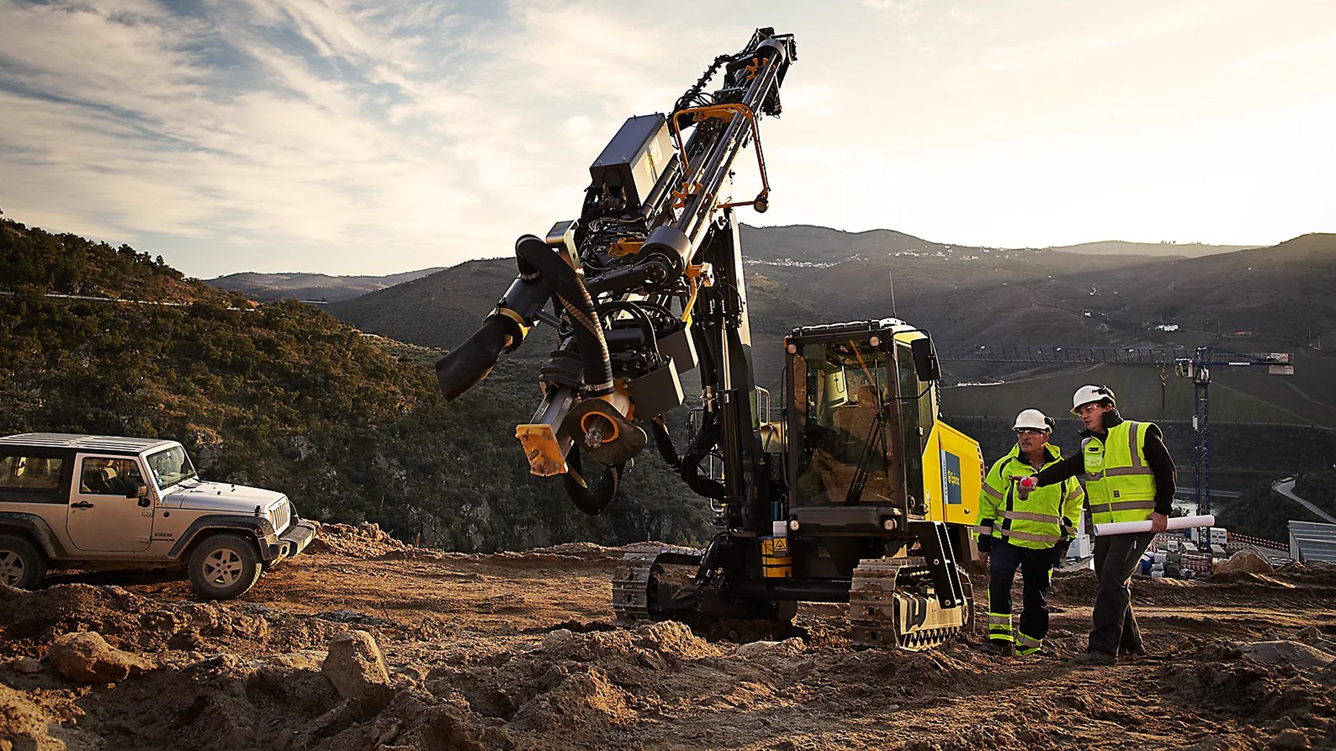 Construction site with a drilling machine, two workers, and a jeep on a dirt road against a mountain landscape.