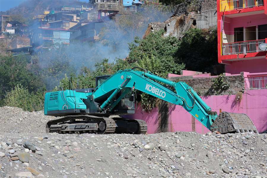 A teal Kobelco excavator on a gravel pile, with smoke and colorful buildings in the background.