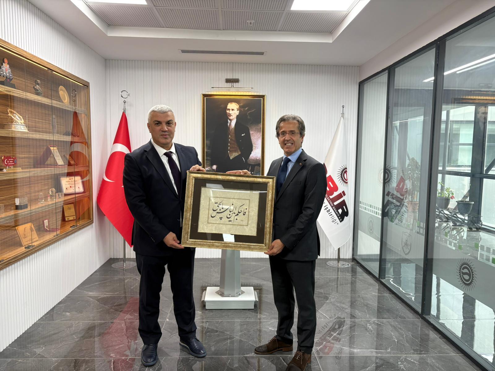 Two men in suits hold a framed artwork in an office, flanked by a Turkish flag and Atatürk portrait.