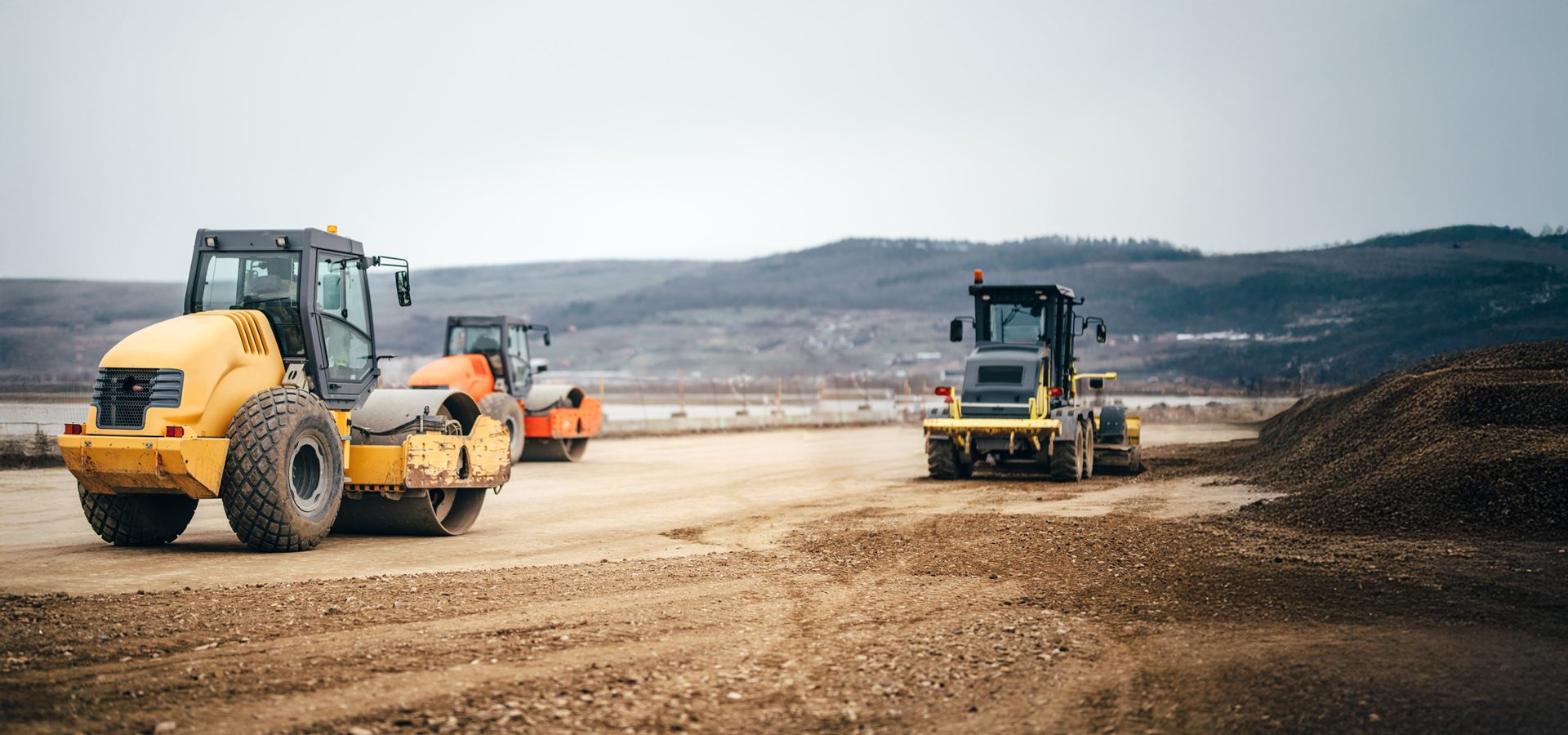 Yellow and orange road rollers and a grader on a dirt construction site with hills in the background.