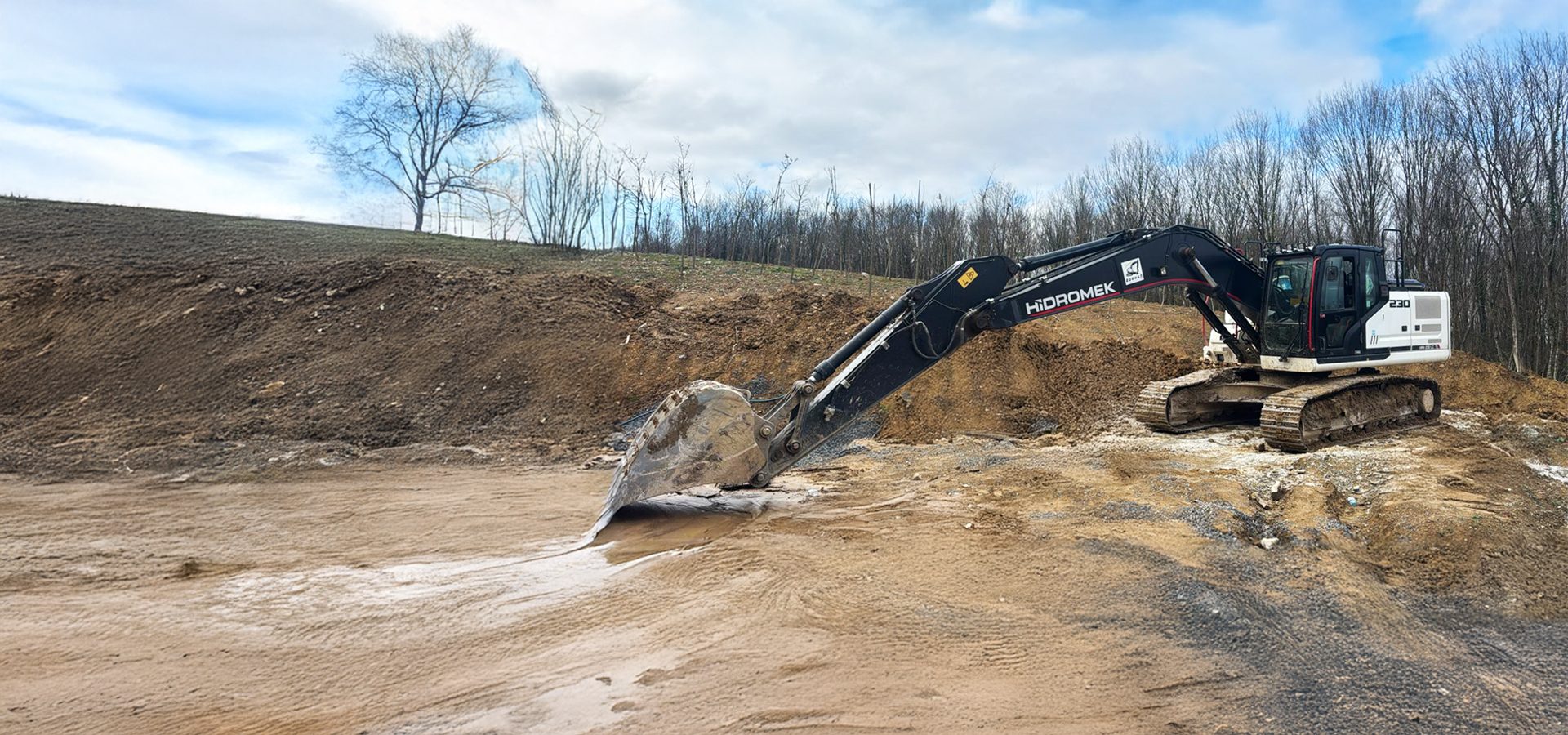 A Hidromek 230 excavator on a muddy construction site, with hills and bare trees under a cloudy sky.