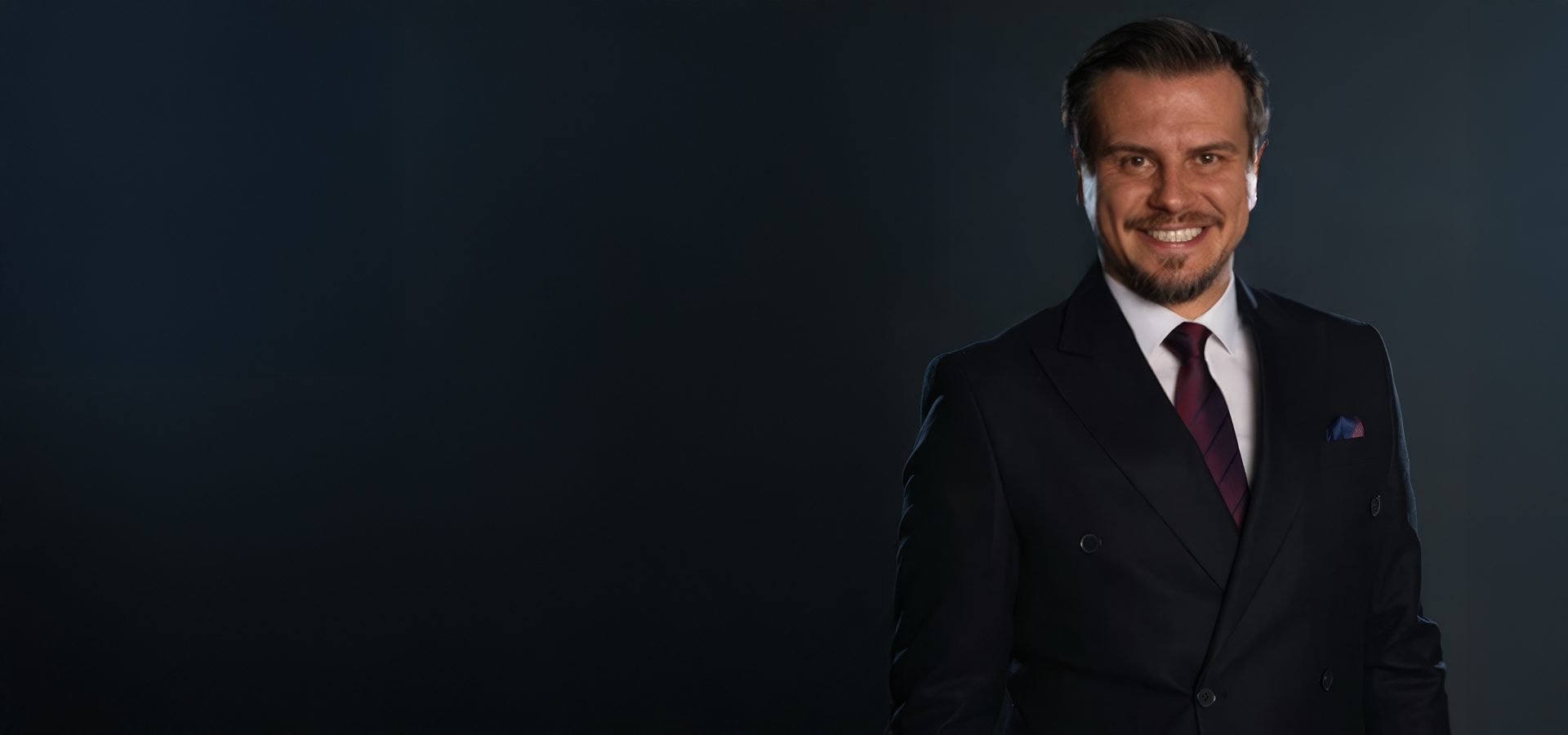 A smiling man in a dark suit and red tie against a dark background.
