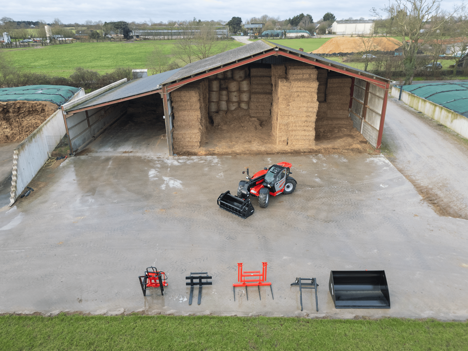 Red telehandler and attachments on a farm, with a large shed full of hay bales in the background.