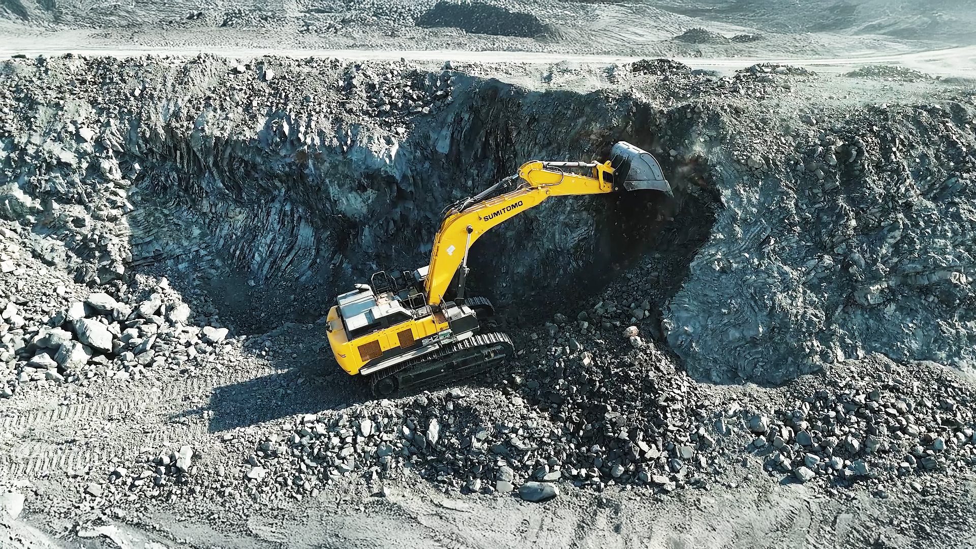 Aerial view of a yellow Sumitomo excavator digging into a steep, rocky quarry wall.