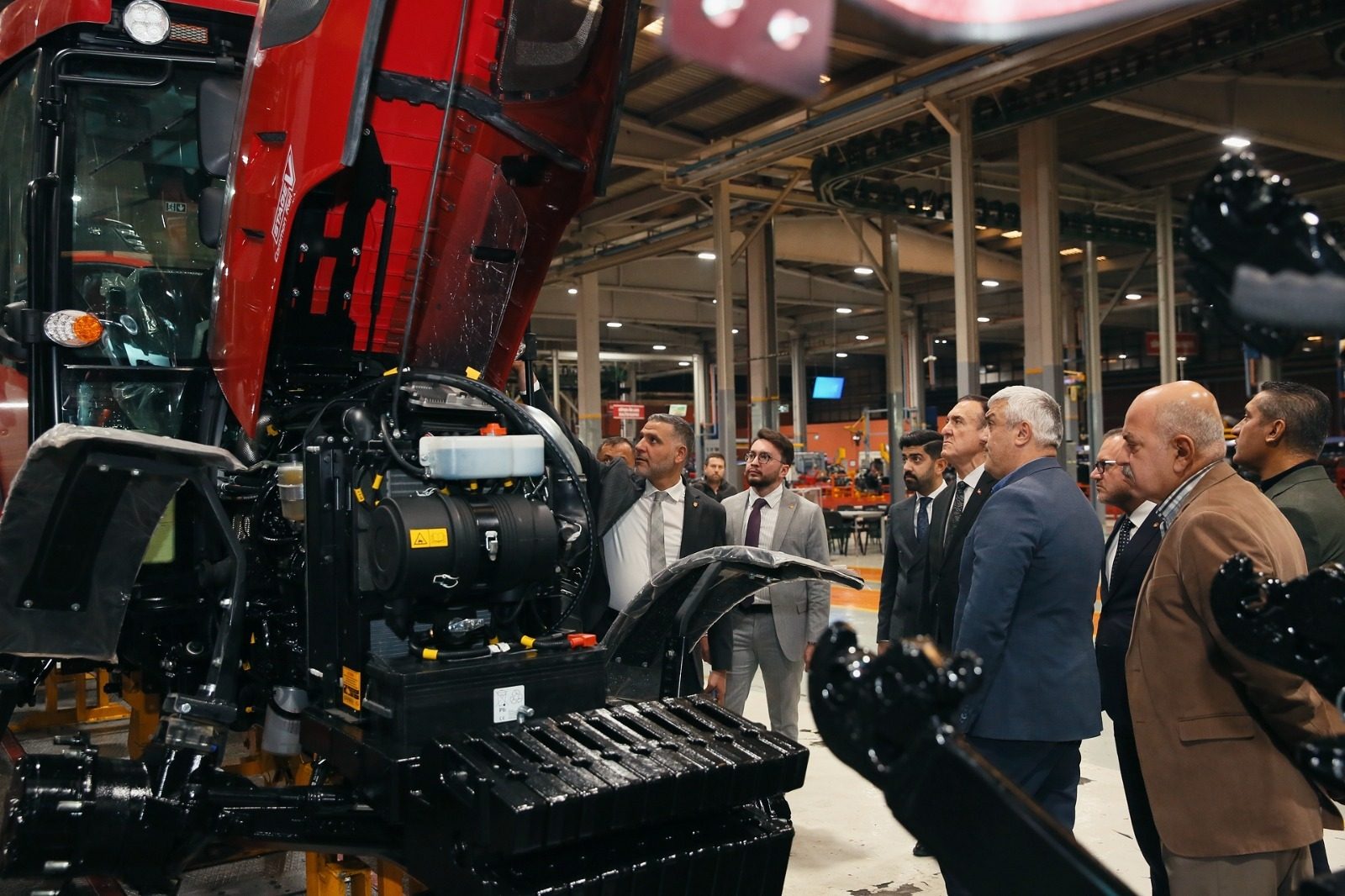 Men in a factory examine the engine of a red tractor with its hood open.